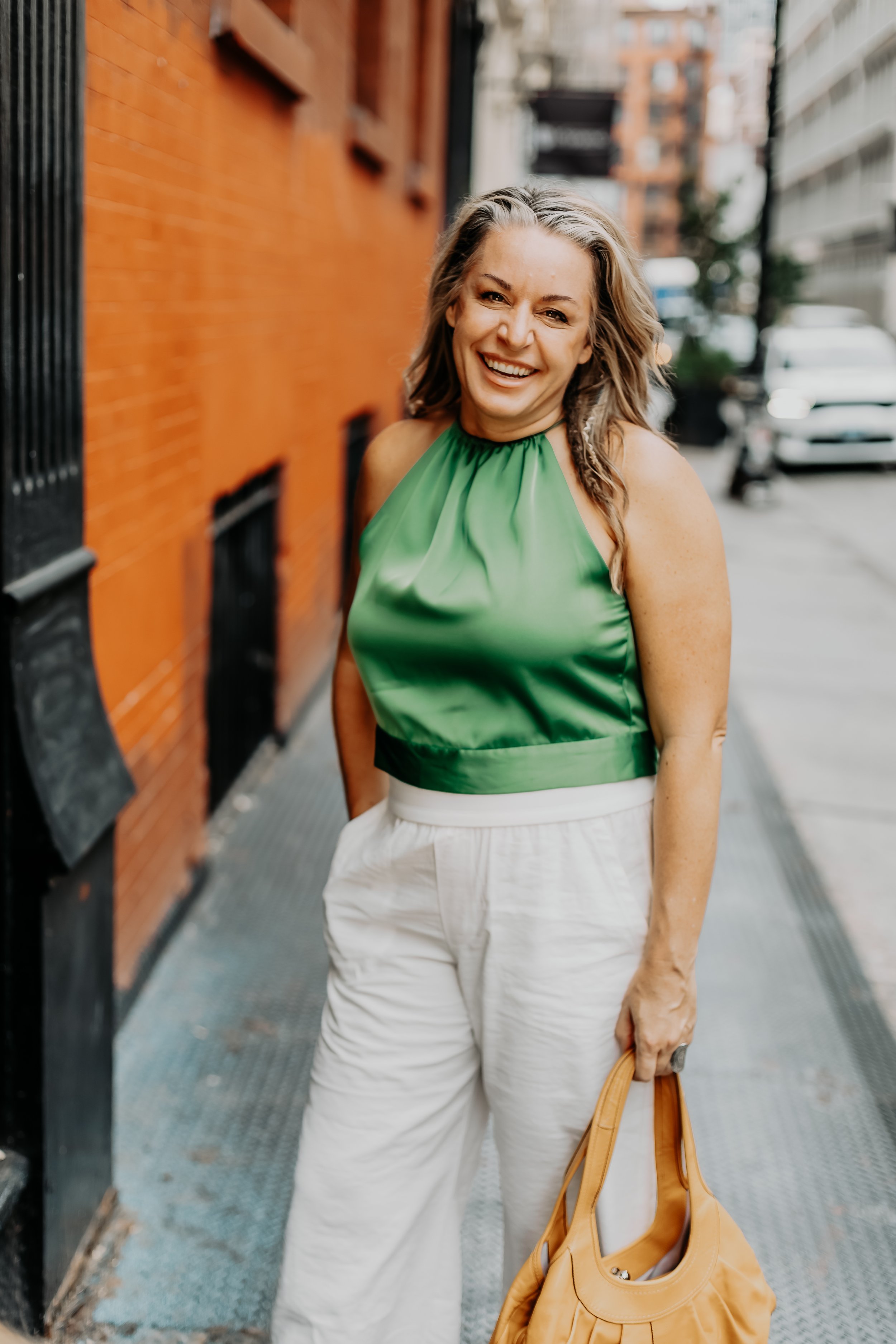 A woman with wavy blonde hair smiling and looking at the camera, standing on an urban street. She is wearing a green halter top and white pants, holding a yellow bag in her right hand. Behind her, there is a red brick building and parked cars.