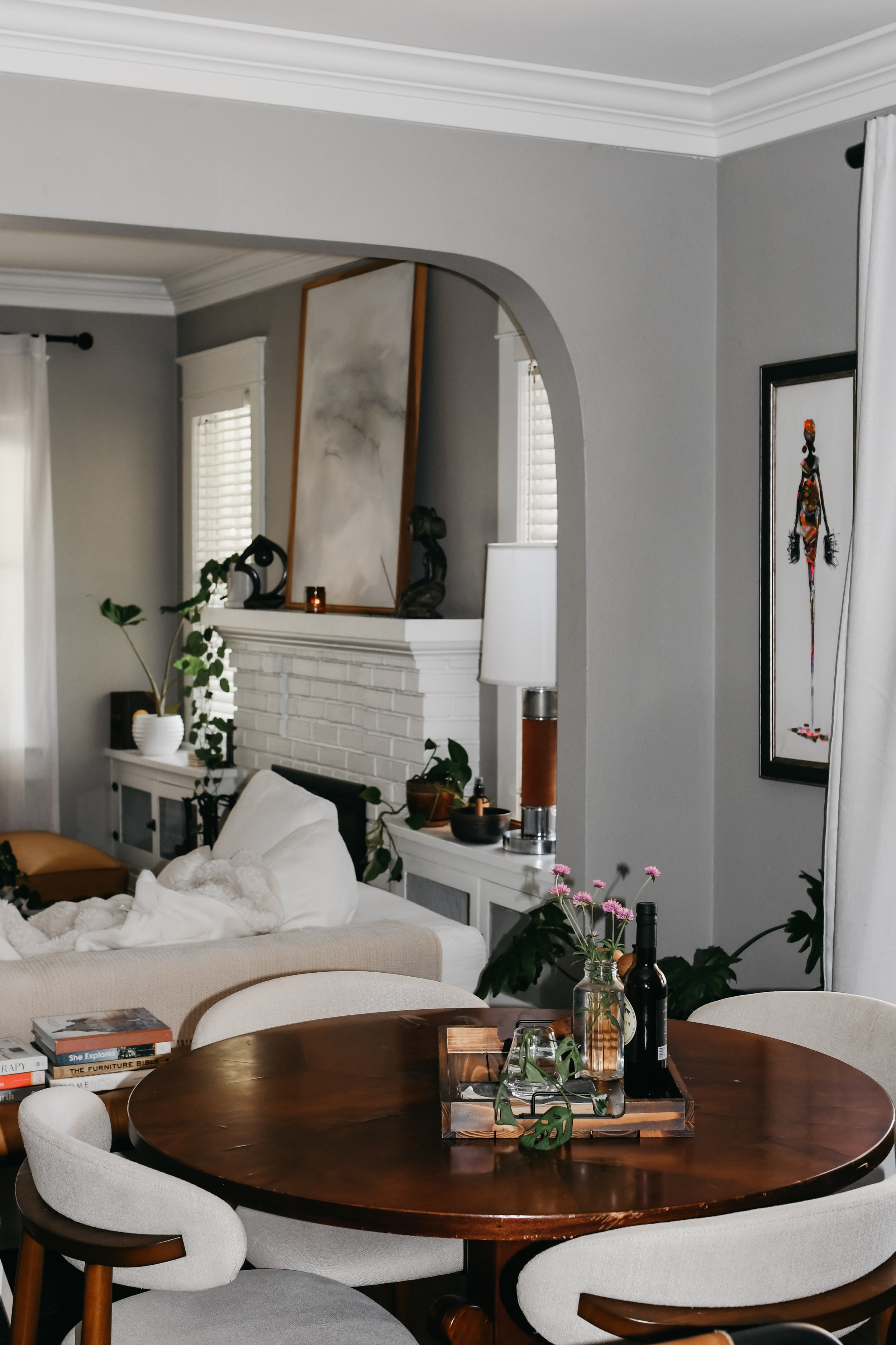 Living room with gray walls, white trim, a fireplace with artwork above, potted plants, and a round wooden dining table with five white and wood chairs, styled with bottles, a tray, and greenery.