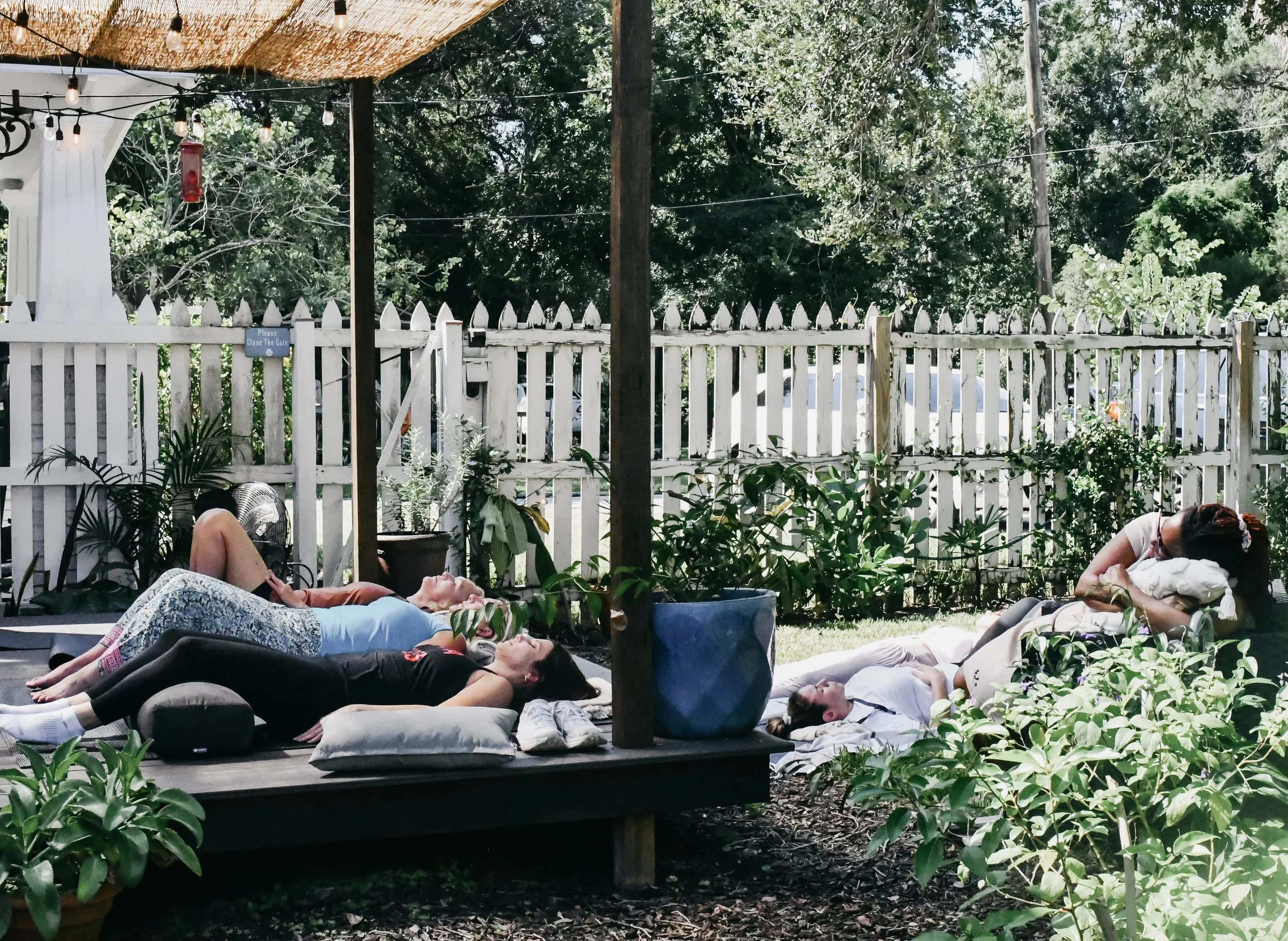 People lying down on cushions and mats, relaxing outdoors on a shaded patio with a white picket fence, plants, and trees in the background.
