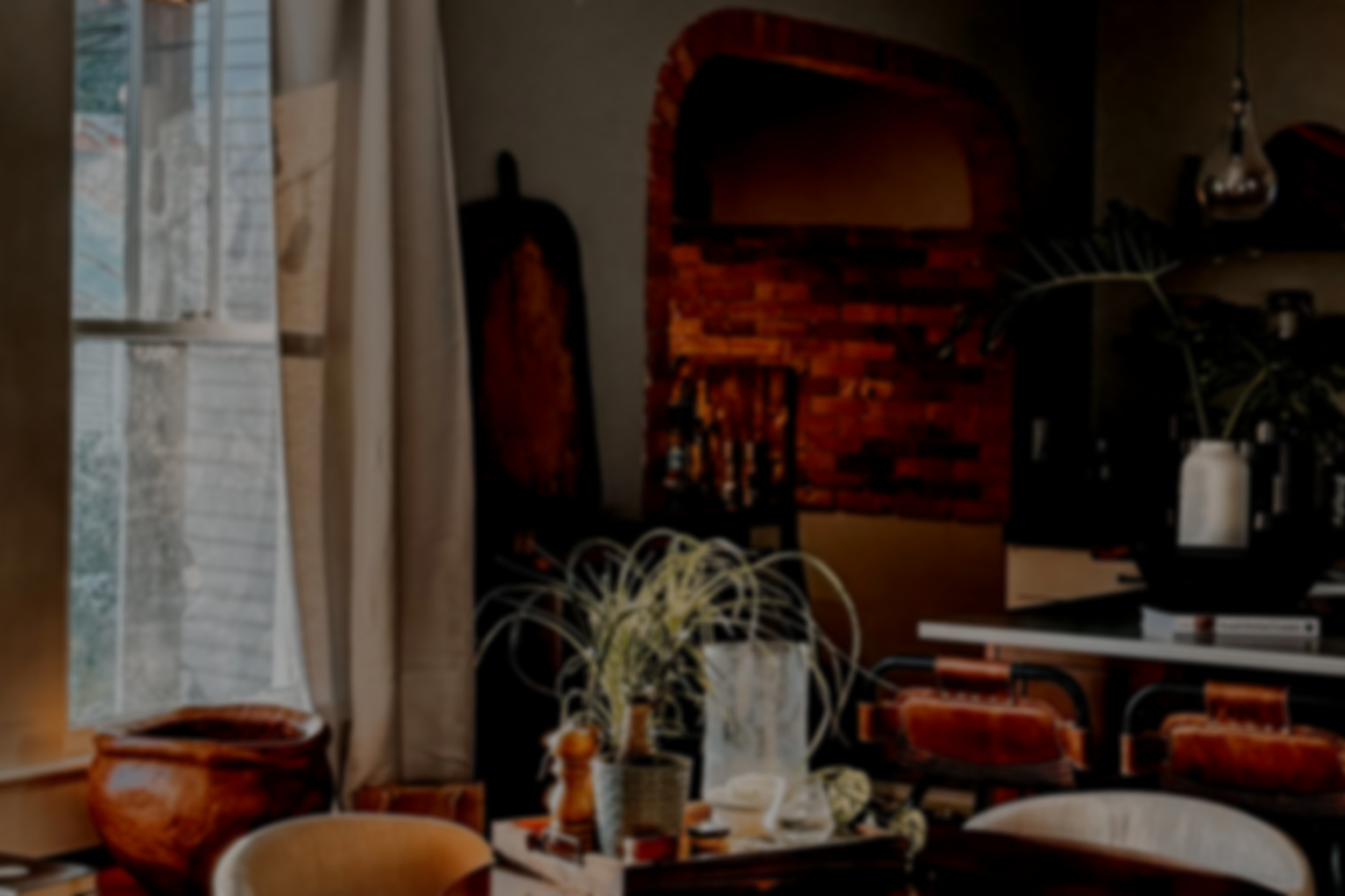A cozy dining area with a table set for a meal, featuring a potted plant centerpiece, surrounded by chairs, with large windows letting in natural light and a brick fireplace in the background.