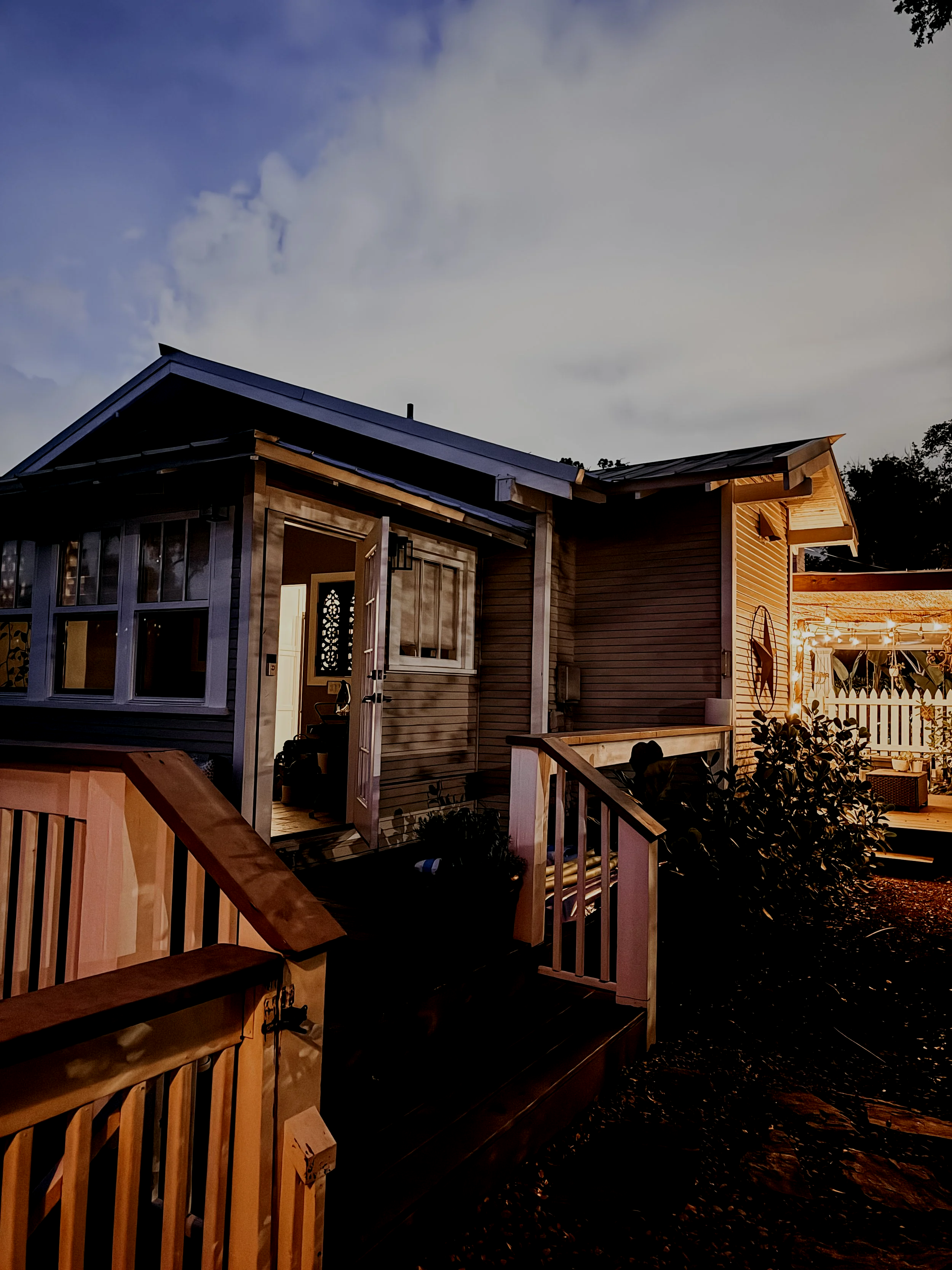 Exterior view of a modern house with large windows, a front porch, and an illuminated backyard patio area at dusk, with a cloudy sky and trees in the background.