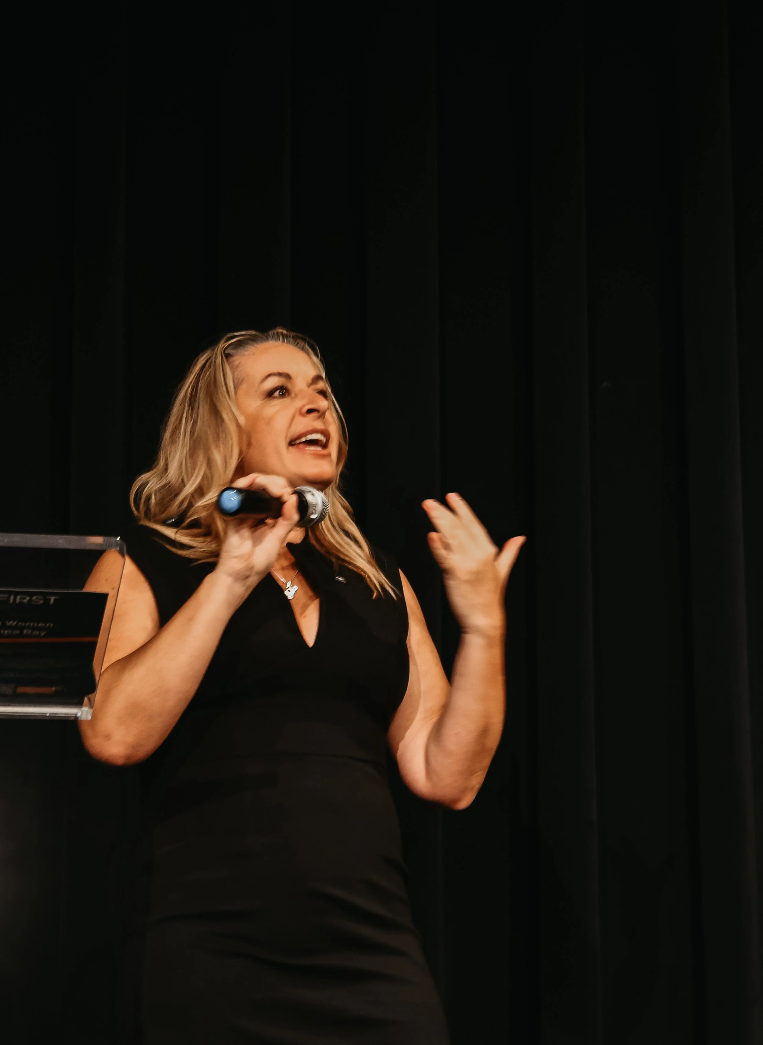 Woman with blonde hair speaking or singing into a microphone on a stage with a black curtain backdrop.