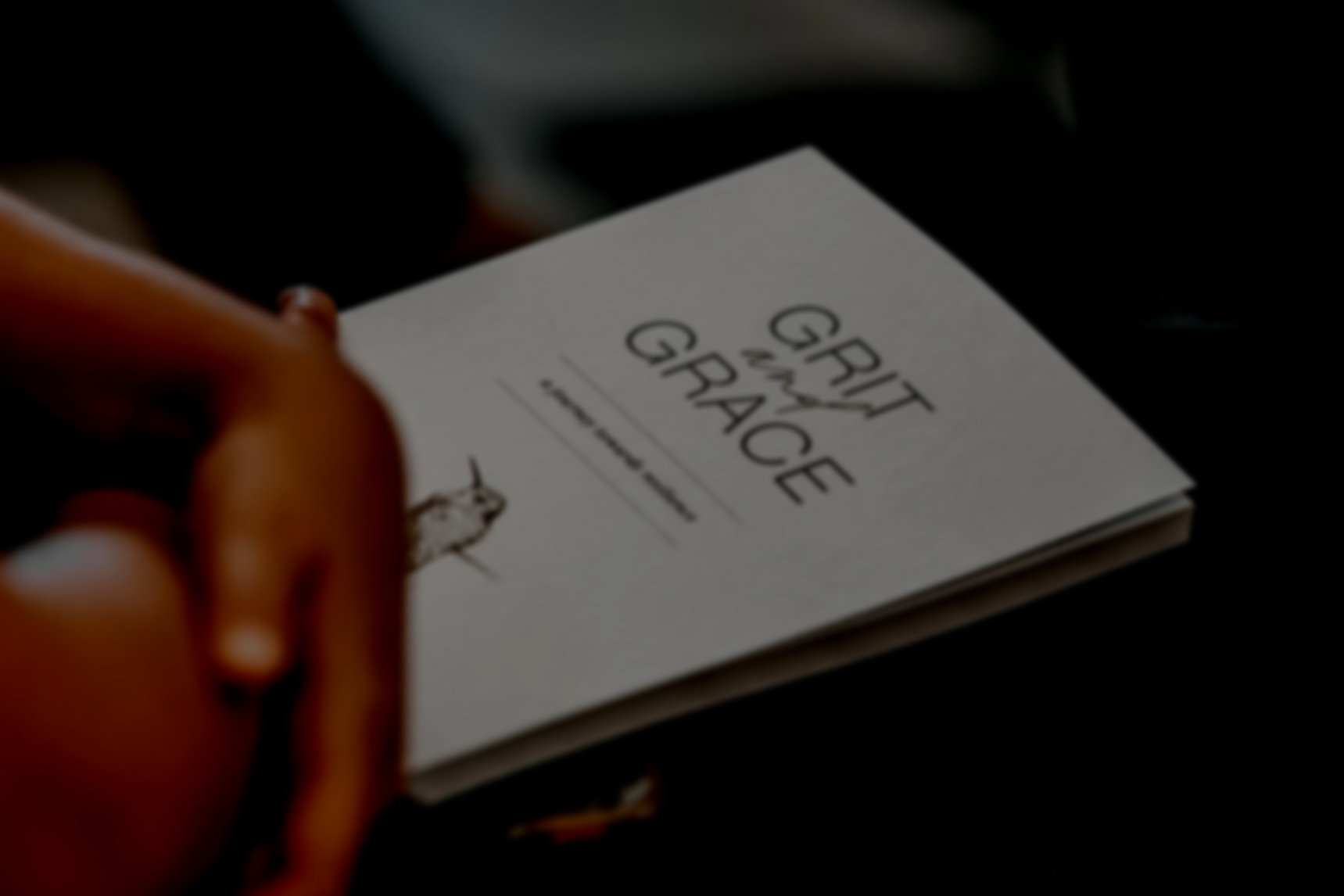 A person holding a book on their lap titled 'Grit and Grace' by Angela Devlen with an illustration of a hummingbird, in a dark setting with the person's fingers visible.