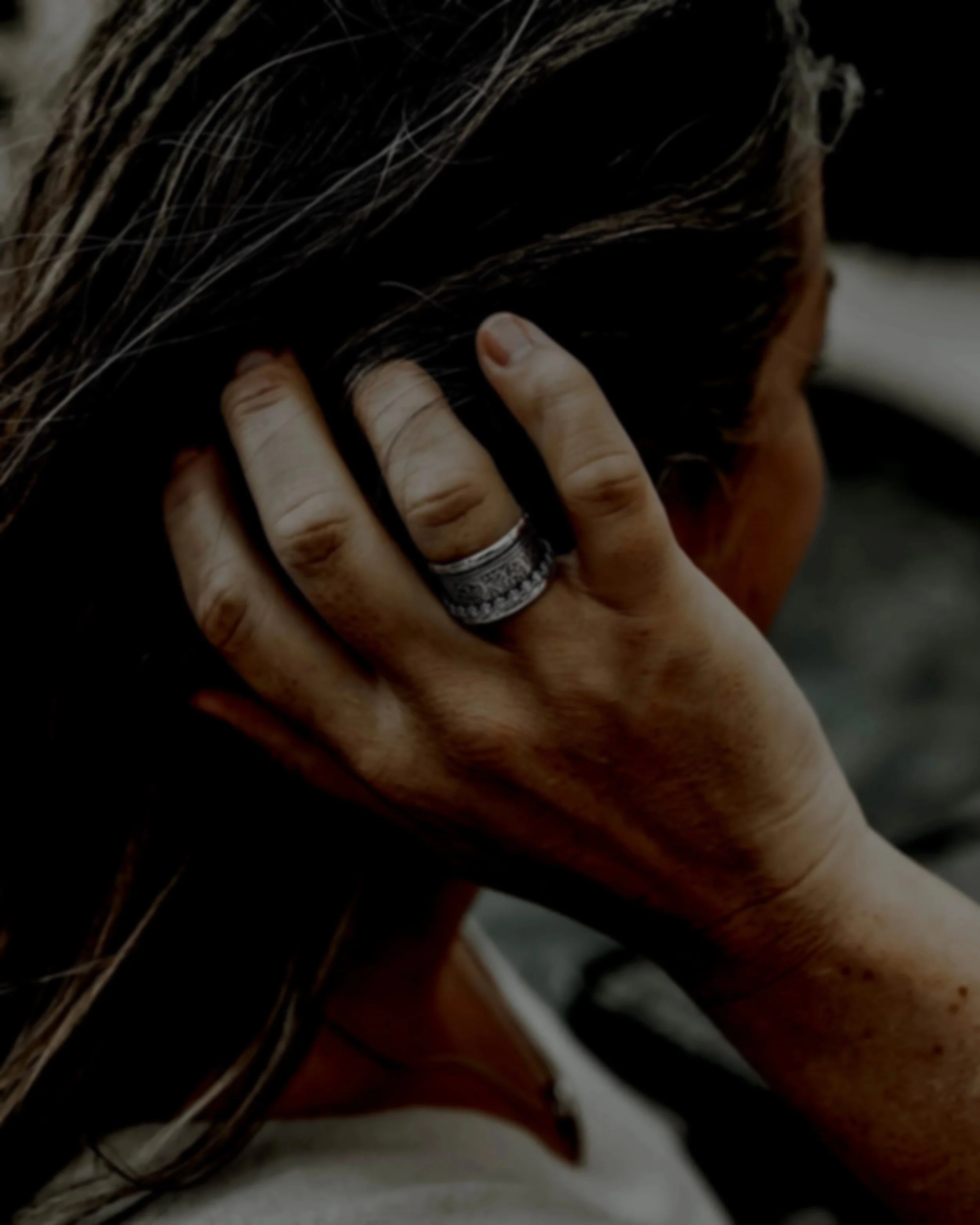 Close-up image of a person with their hand on their head, fingers resting on dark, layered hair. The person is wearing a prominent silver ring on their middle finger.