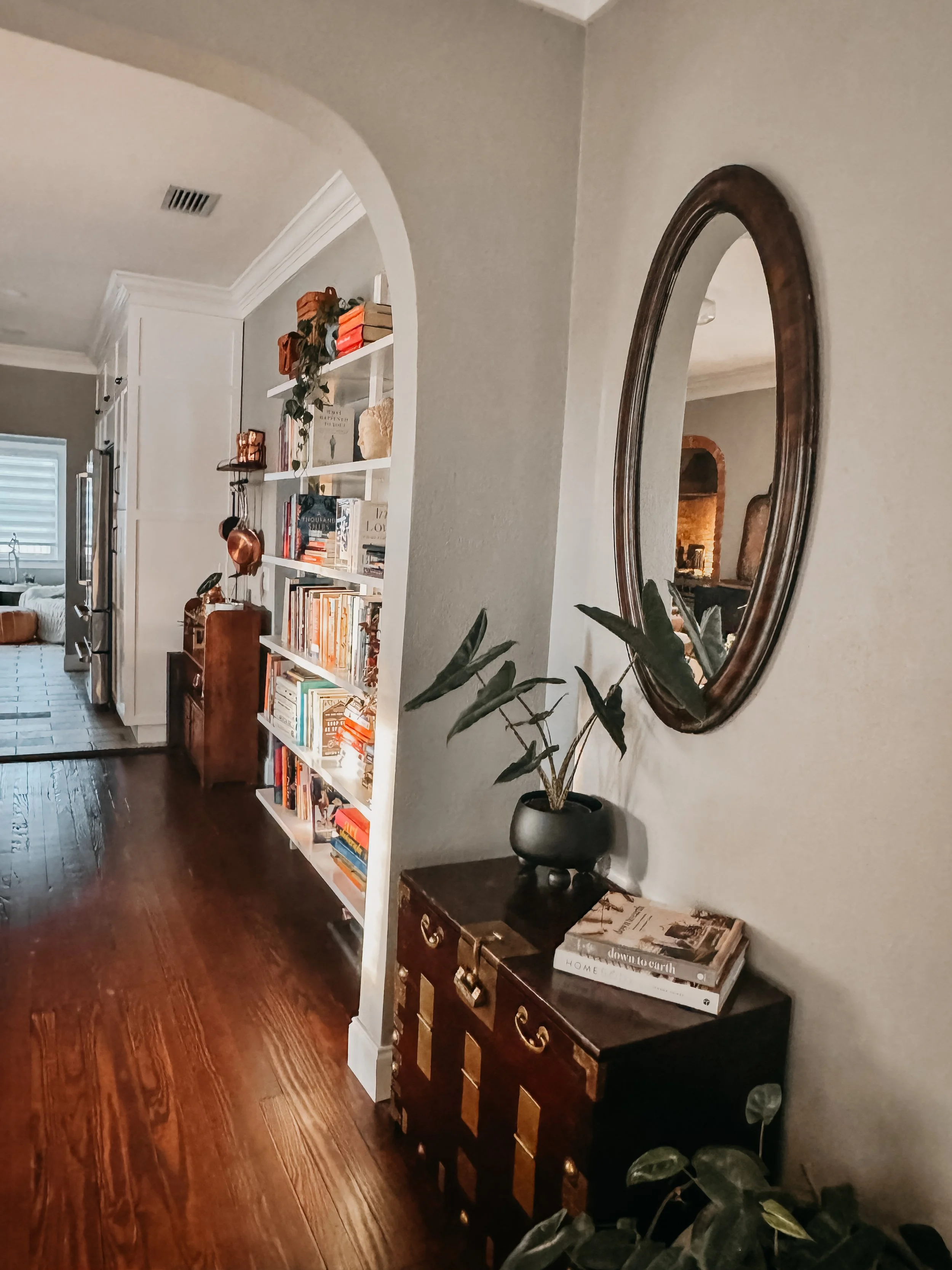A cozy living room with a large white bookshelf filled with books and decorative items, a wooden side table with magazines, a potted plant, and a large oval mirror on the wall reflecting part of the room.