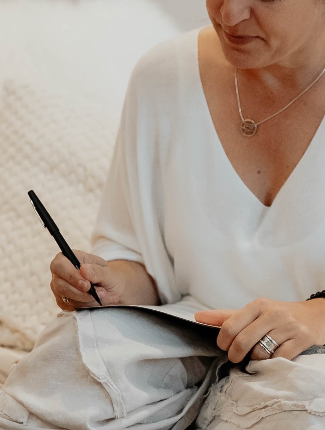 A woman wearing a white top and beige pants, sitting while writing in a notebook with a black pen. She has a necklace and a ring on her finger.