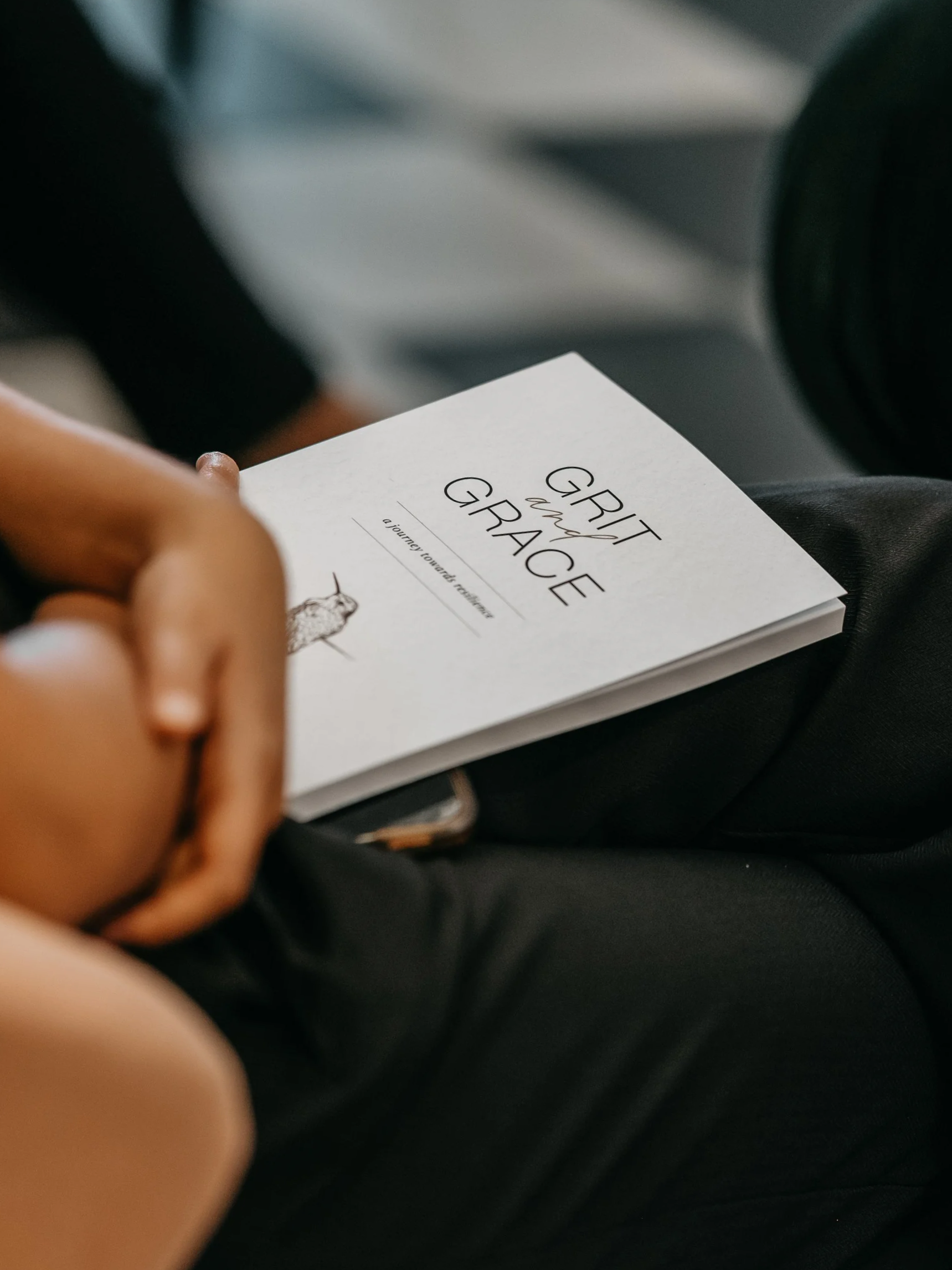 Close-up of a person holding a white card titled 'GRIT and GRACE' with the subtitle 'a journey towards resilience' in a car seat.