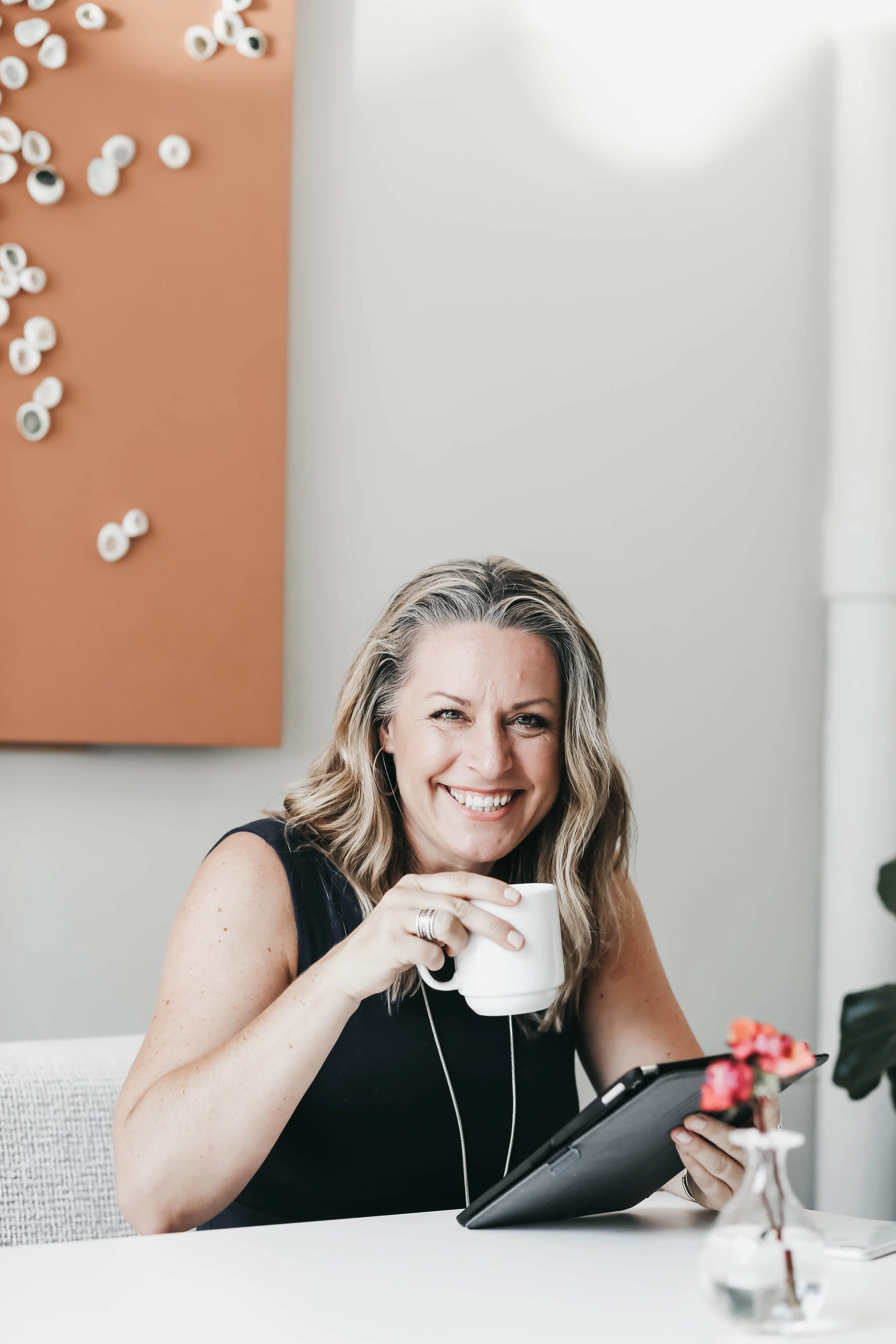 A woman with wavy blonde hair and earrings, smiling while holding a white mug in her right hand and a tablet in her left hand, seated at a white table in a room with a white and orange wall and small white decorations on the wall.