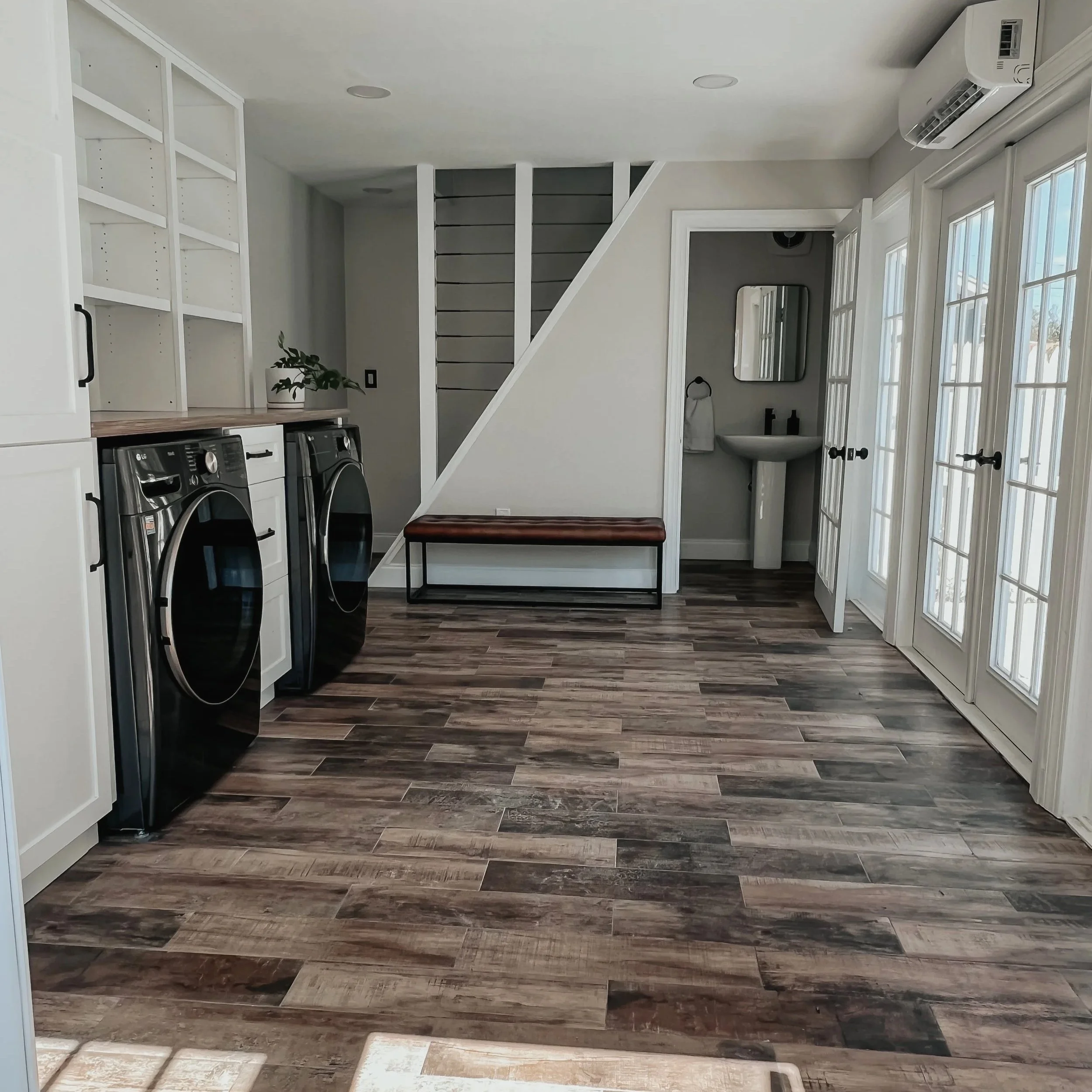 Interior of a modern laundry room with wood-like tile flooring, white cabinets, front-loading washer and dryer, small mirror, and a door with glass panels leading outside.