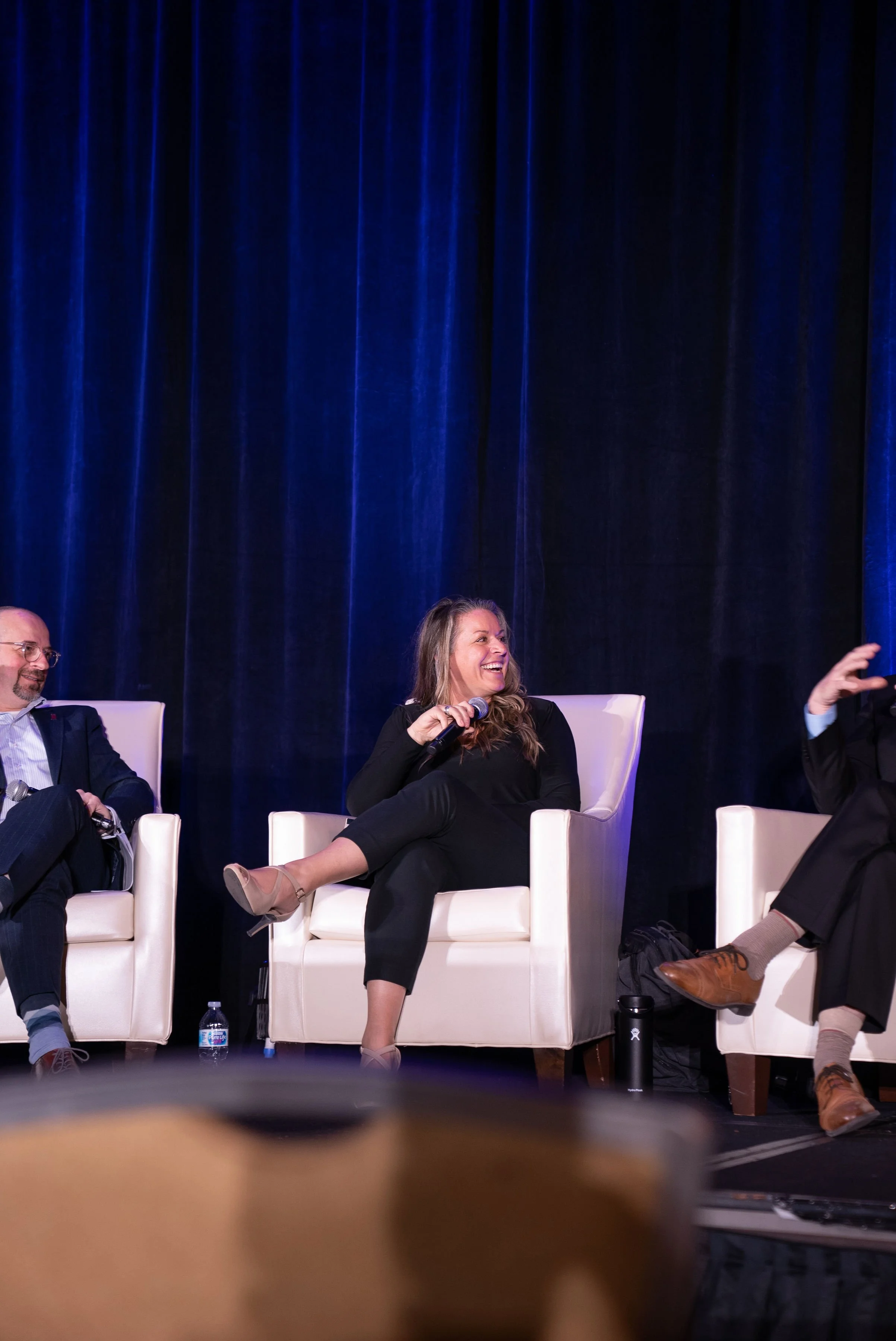 People sitting on stage chairs during a panel discussion, with a woman in the center smiling and holding a microphone, in front of a blue curtain.