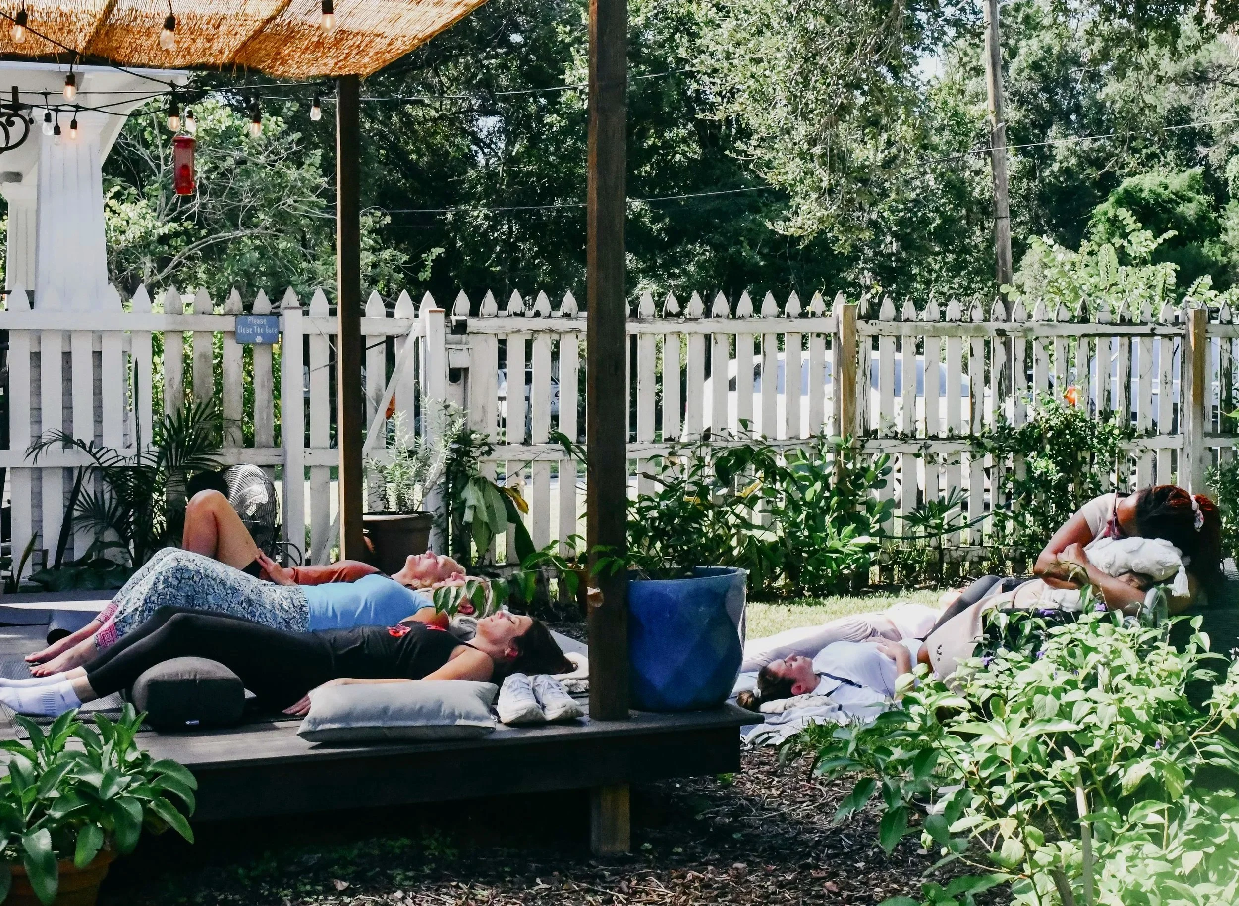 People lying down and relaxing on a shaded outdoor deck with cushions, surrounded by green plants and a white picket fence.