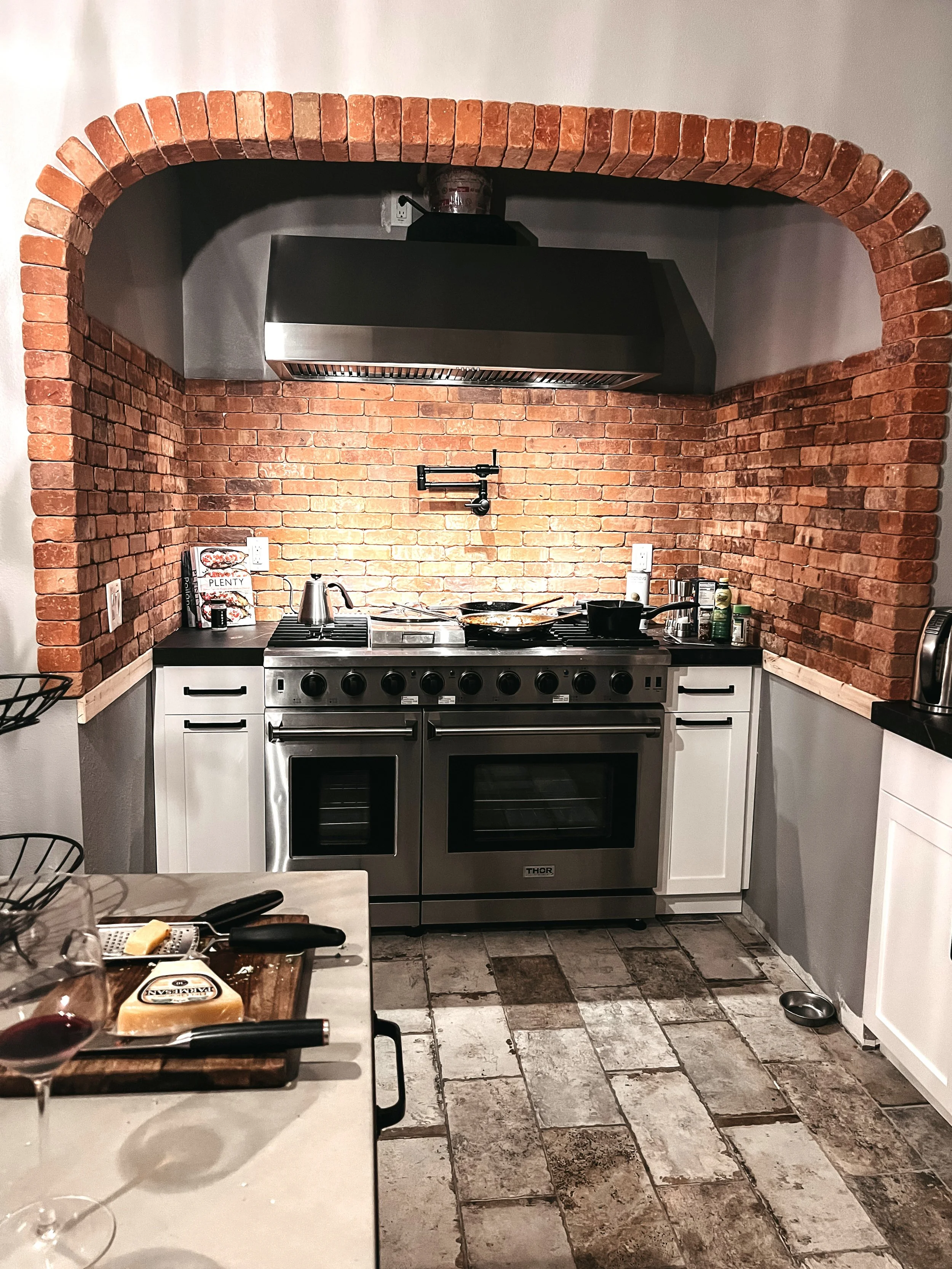A modern kitchen with a brick archway, stainless steel stove and oven, and white cabinets. There are granite countertops with a cheese block, a cheese knife, wine glasses, and a cutting board in the foreground.