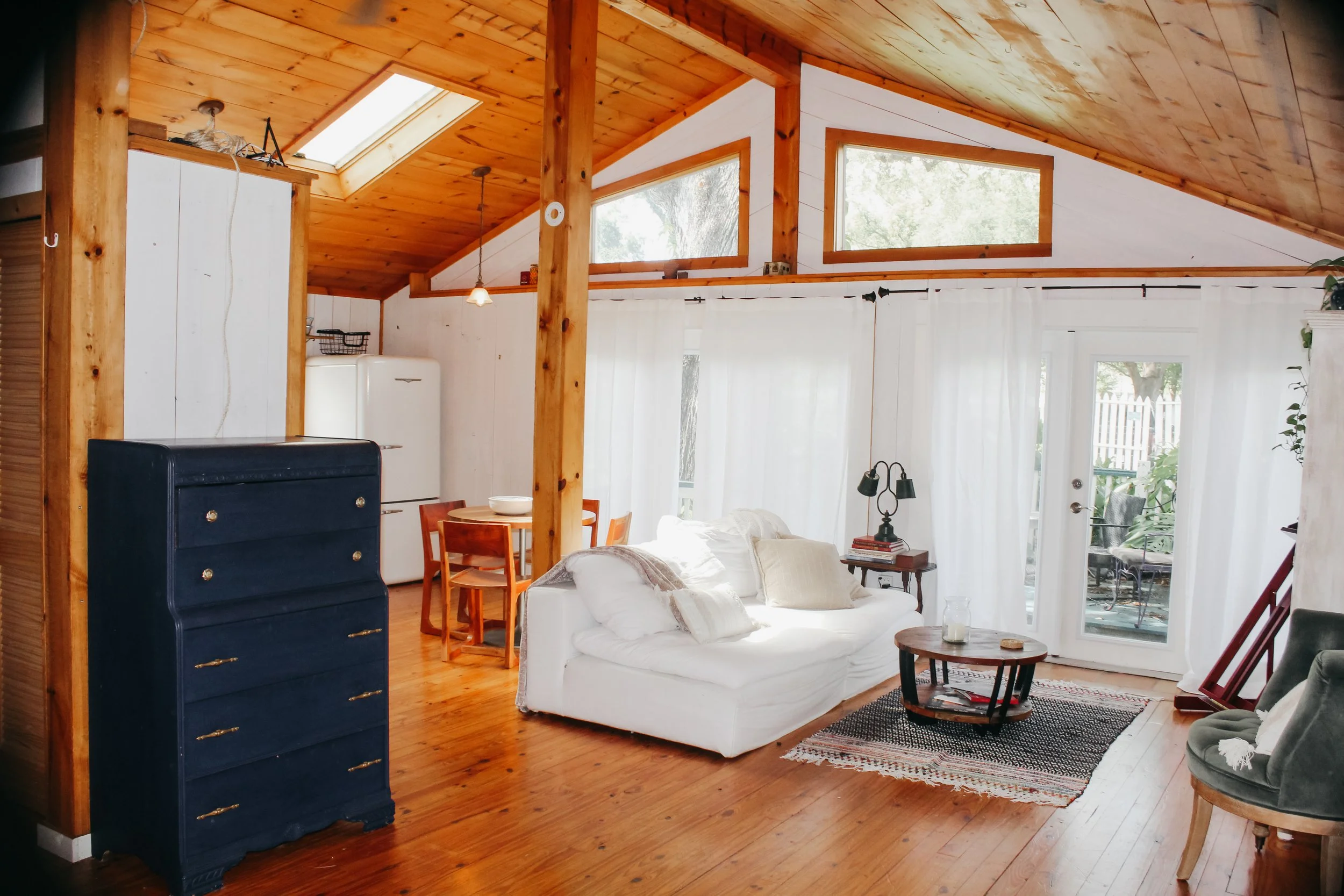 Interior view of a cozy living space with wooden ceiling and floors, white walls, large windows, white sofa, small round coffee table, blue dresser, and dining area with a small wooden table and chairs.