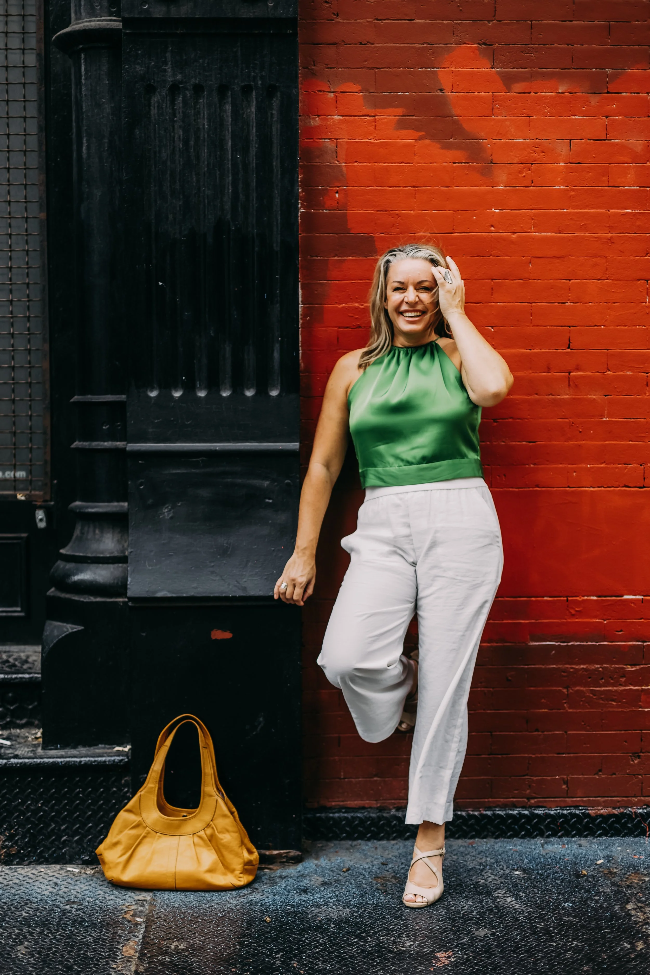 A woman with blonde hair, wearing a green sleeveless top and white pants, is laughing and touching her hair while standing against a red brick wall. There is a large yellow handbag on the ground beside her.