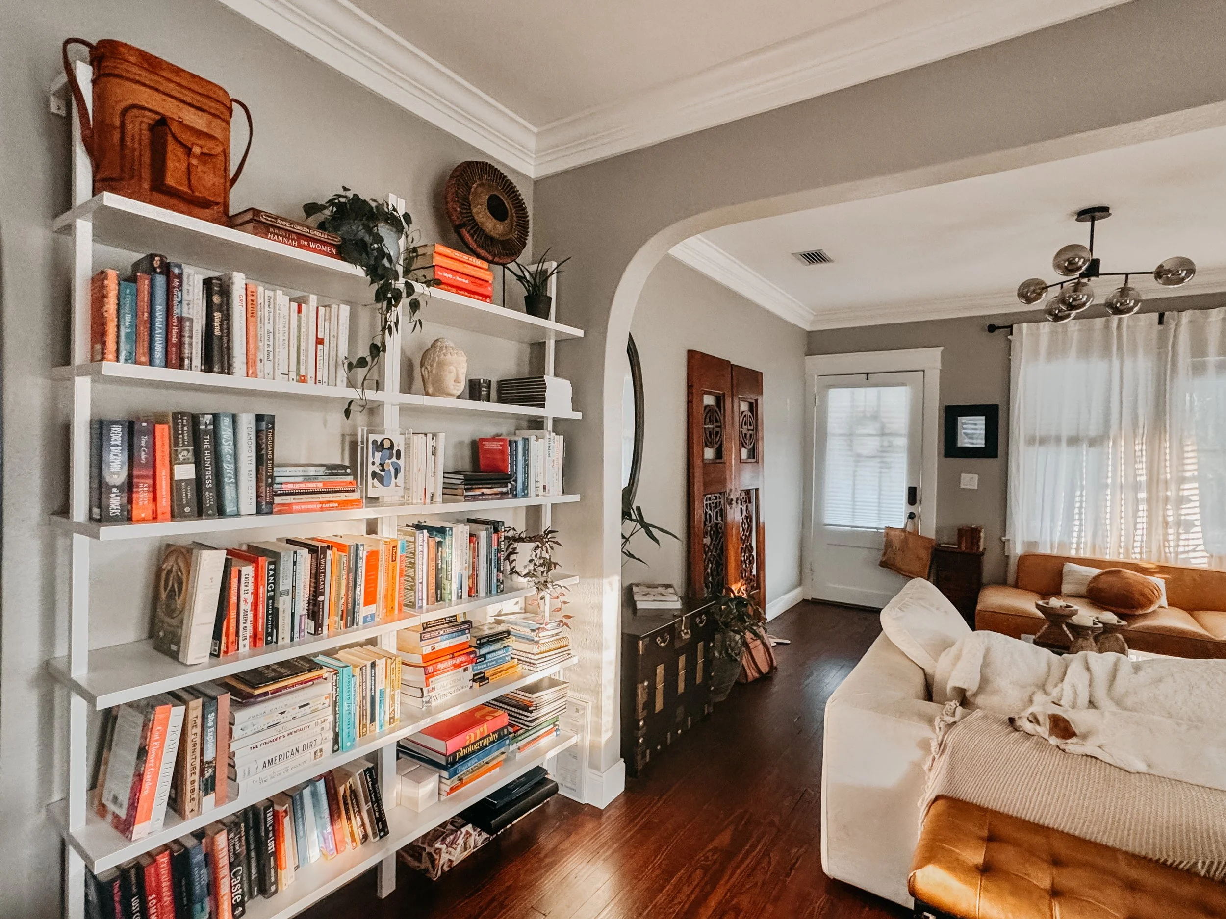 A living room with a white bookshelf filled with books, some decorative items, and plants. There is a doorway with a window and curtains, a sofa with pillows, and a modern ceiling light fixture.
