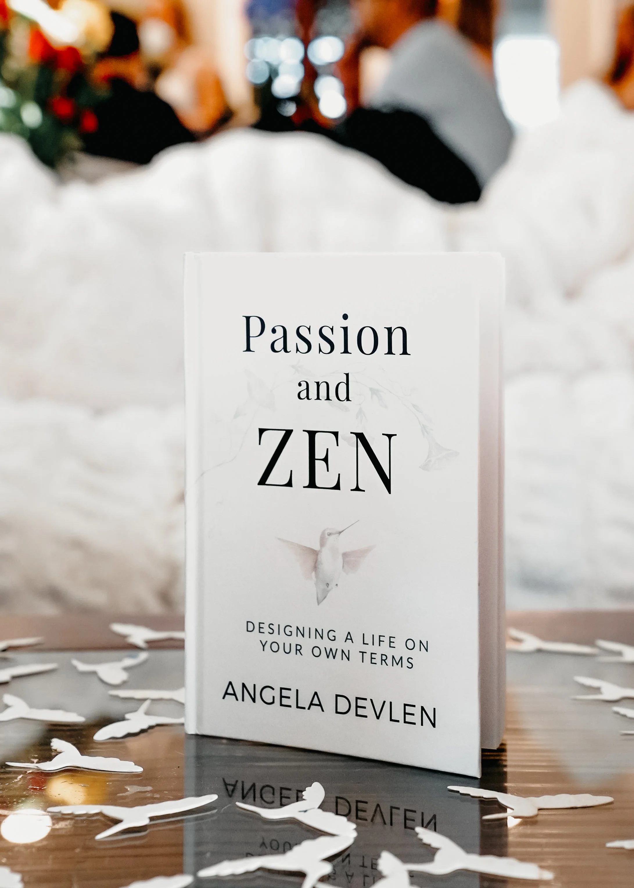 A book titled 'Passion and Zen' by Angela Devlin stands on a table decorated with scattered paper cutouts shaped like birds. In the background, blurred festive holiday decorations and people are visible.