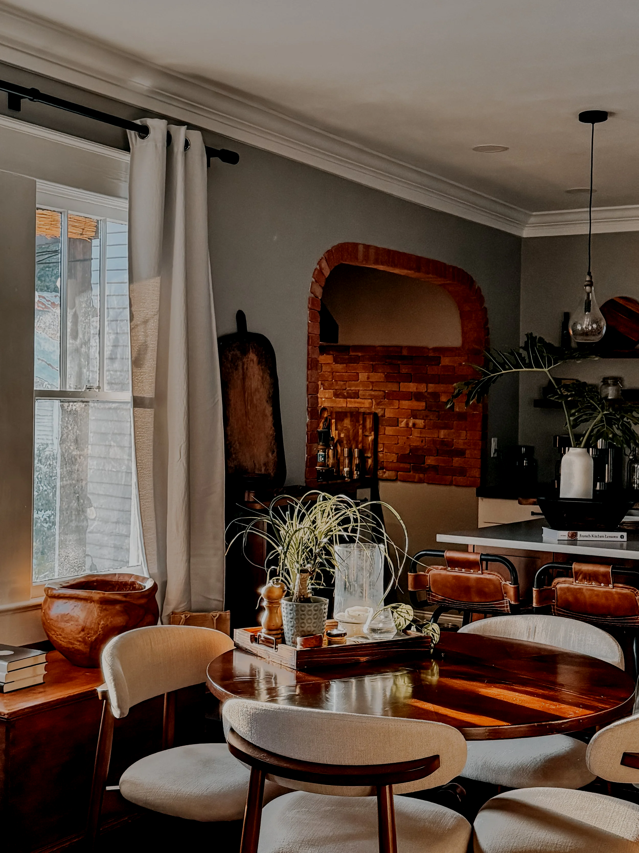 A dining area with a round wooden table surrounded by beige upholstered chairs. Plants and decorative items are on the table. A brick archway and a fireplace are visible in the background, along with a window with curtains.