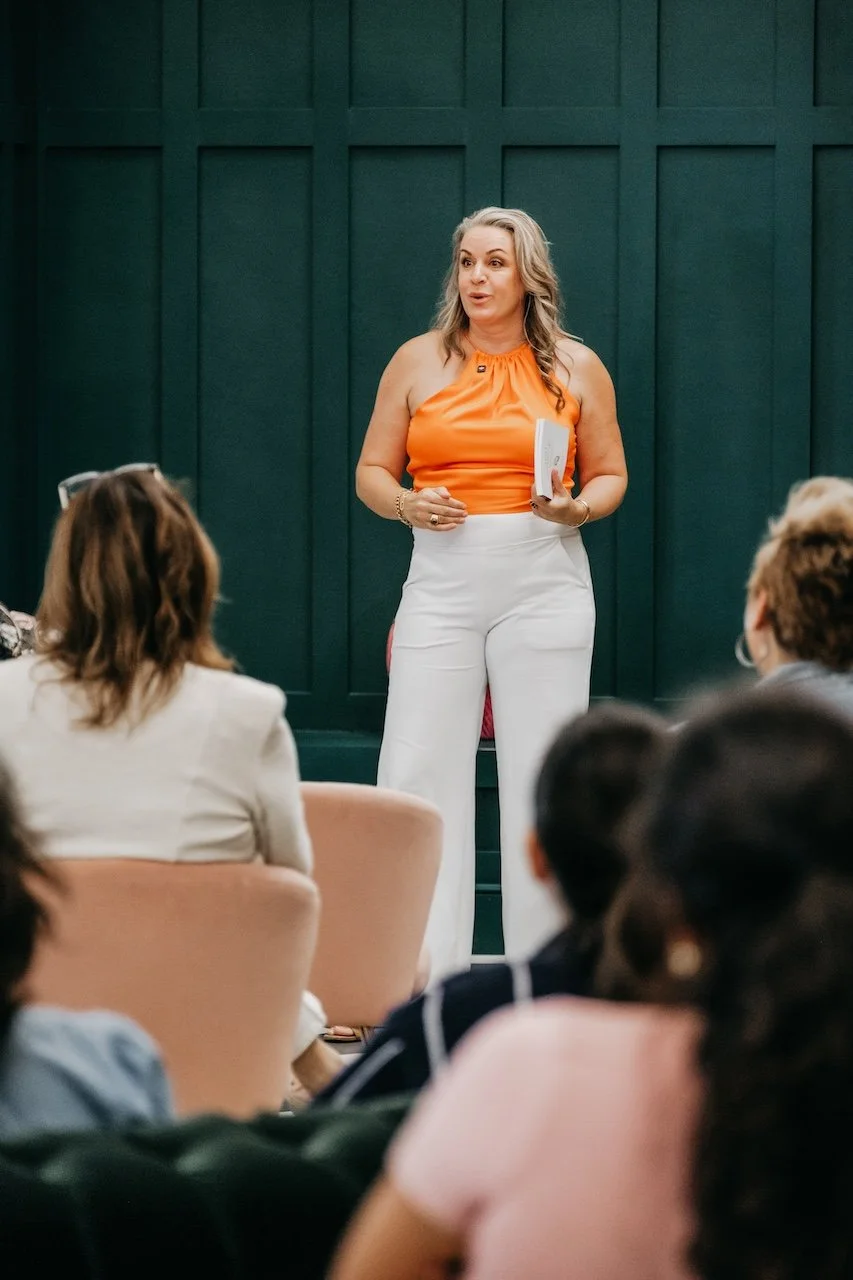 A woman in an orange sleeveless top and white pants giving a presentation to an audience in a room with dark green paneled walls.