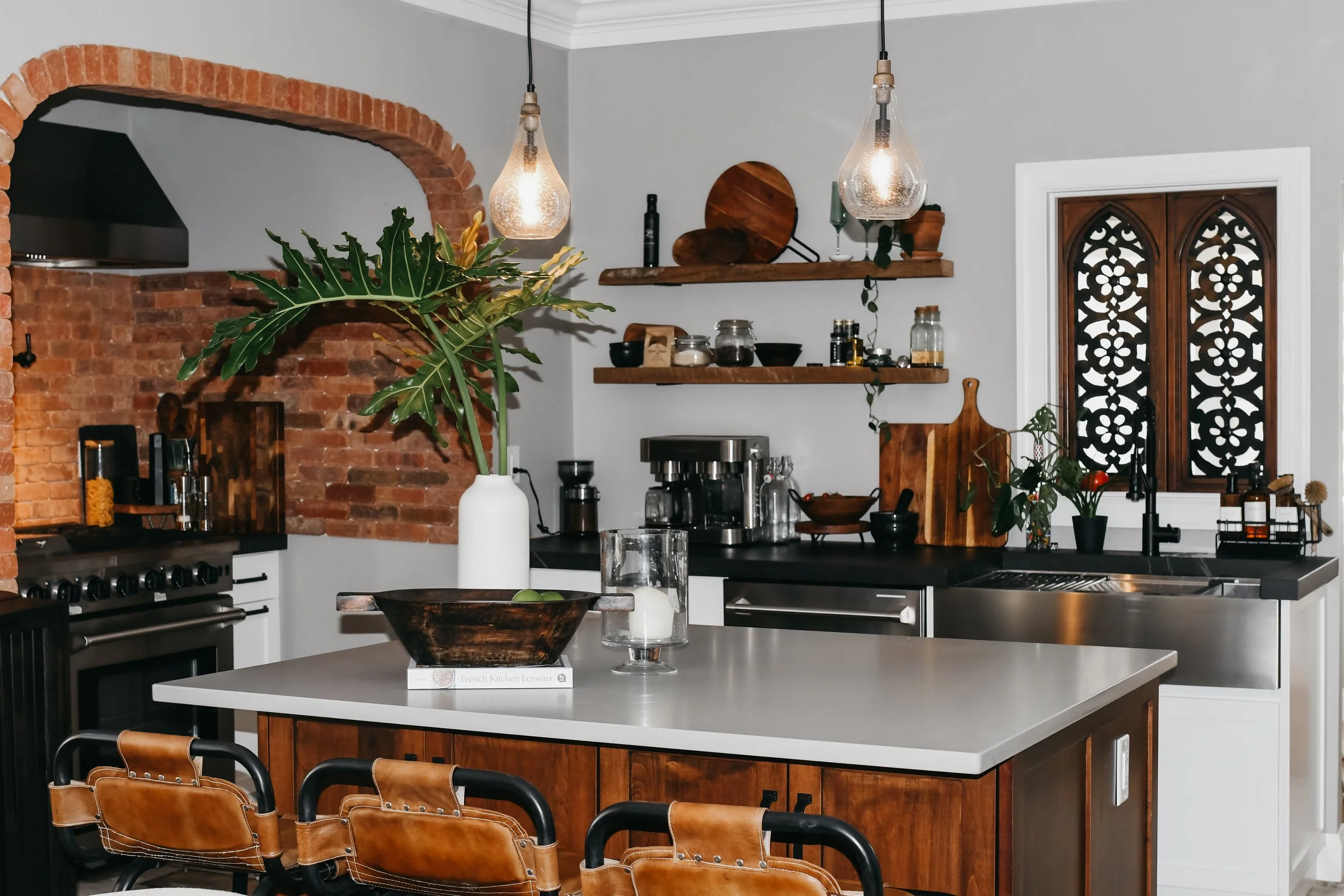 A modern kitchen with exposed brick archway, wood floating shelves, and decorative elements. There are pendant lights hanging over a white kitchen island, which has a plant in a white vase, a bowl with green apples, and a glass of water. The background includes kitchen appliances and a window with decorative wooden panels.
