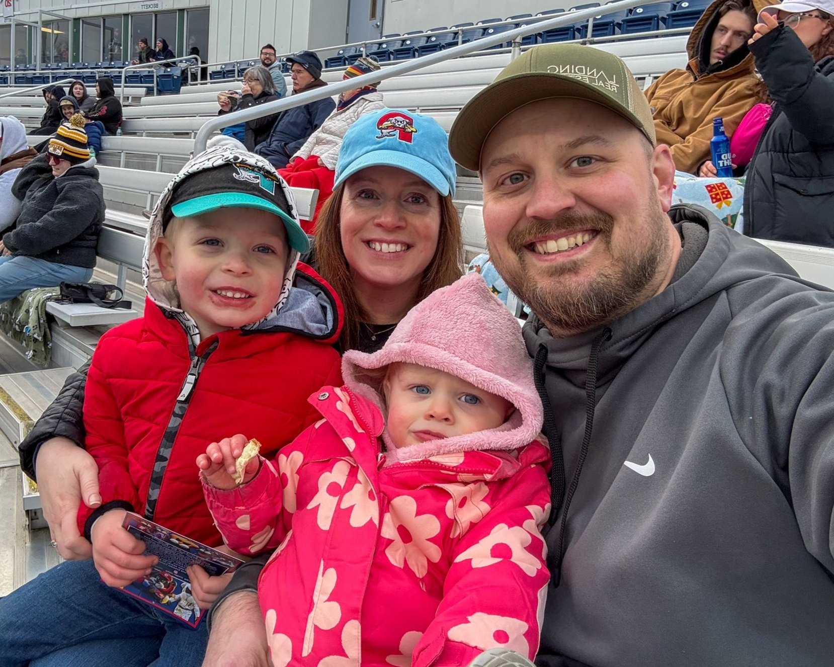 Joe Auger, Joe Auger Real Estate, Joe Auger Maine, A family of four smiling for a selfie at a stadium, with other spectators sitting in the background.