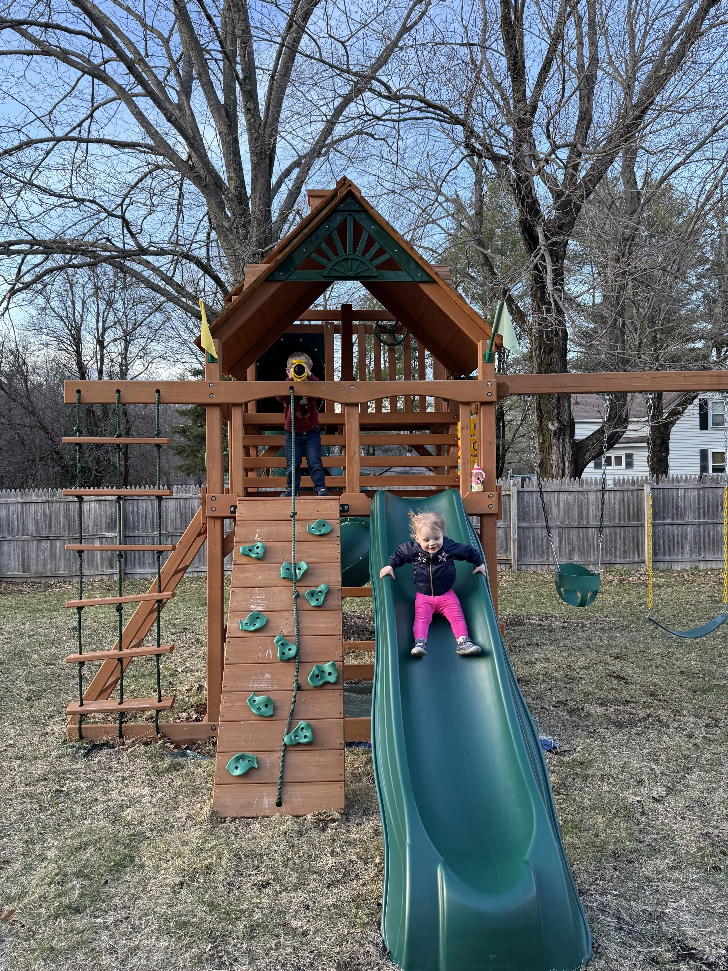 Joe Auger, Joe Auger Real Estate, Joe Auger Maine, Children playing on a wooden backyard playset that has a slide, climbing wall, swings, and a small house at the top, with leafless trees and a wooden fence in the background.