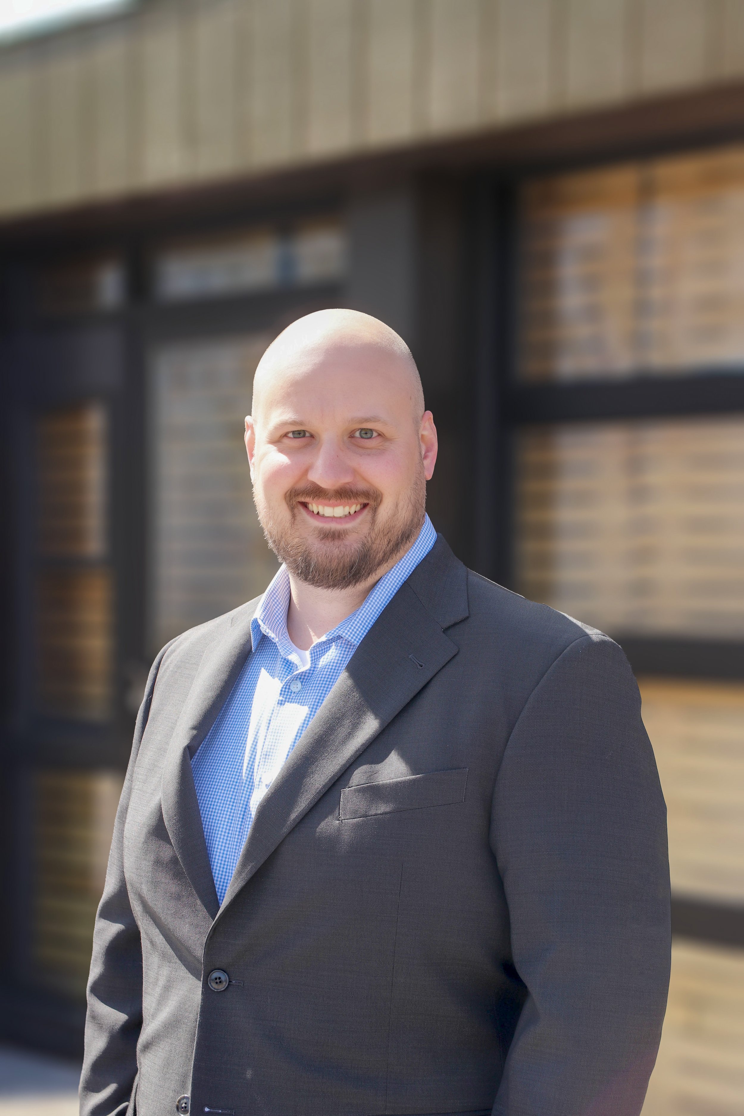 Joe Auger, Joe Auger Real Estate, Joe Auger Maine, A smiling man with a beard and shaved head wearing a dark blazer and light blue dress shirt, standing outdoors in front of a wooden building.