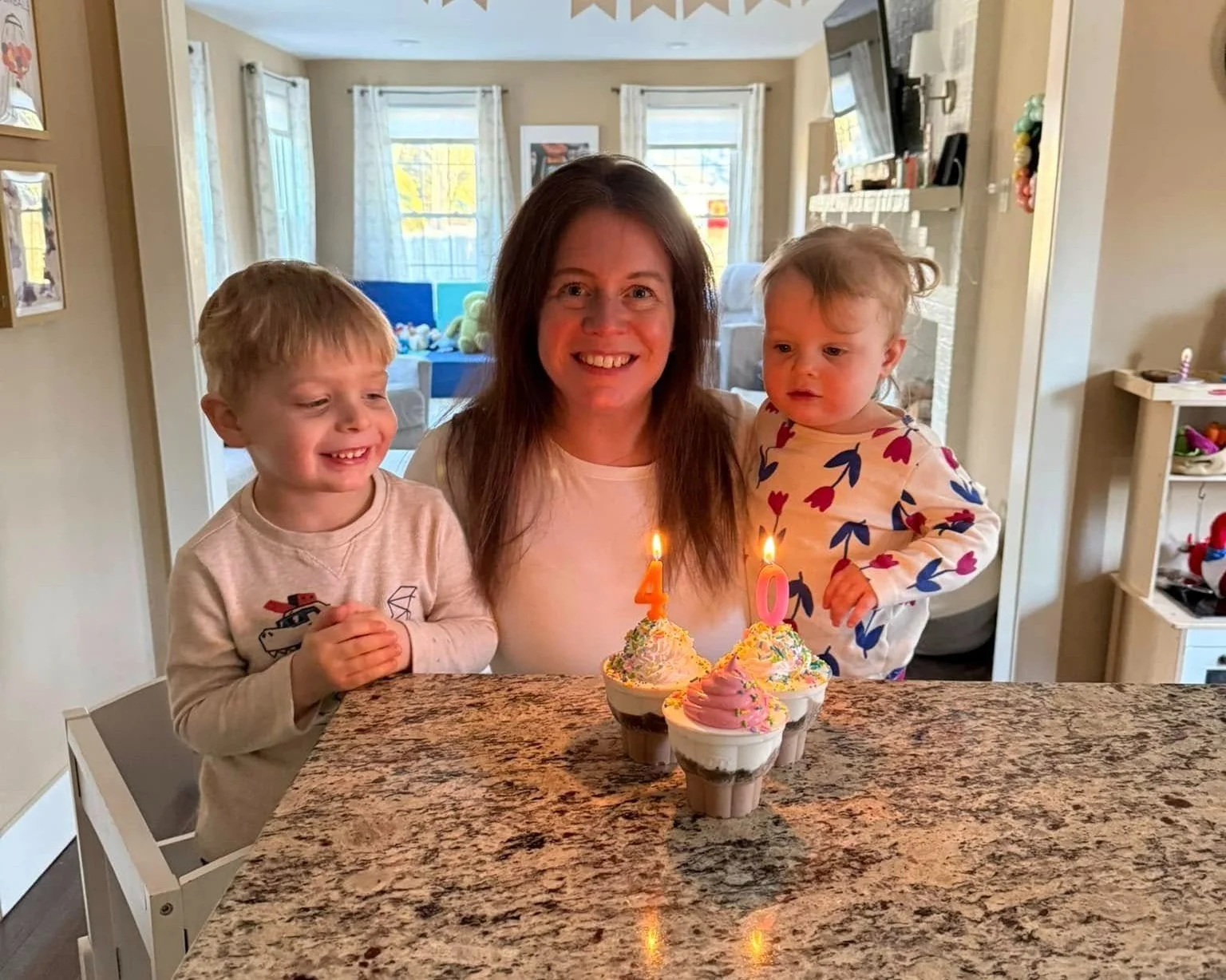 Joe Auger, Joe Auger Real Estate, Joe Auger Maine, A woman with two young children celebrating a birthday with cupcakes on a kitchen counter, candles on the cupcakes, and a smiling woman in the center.
