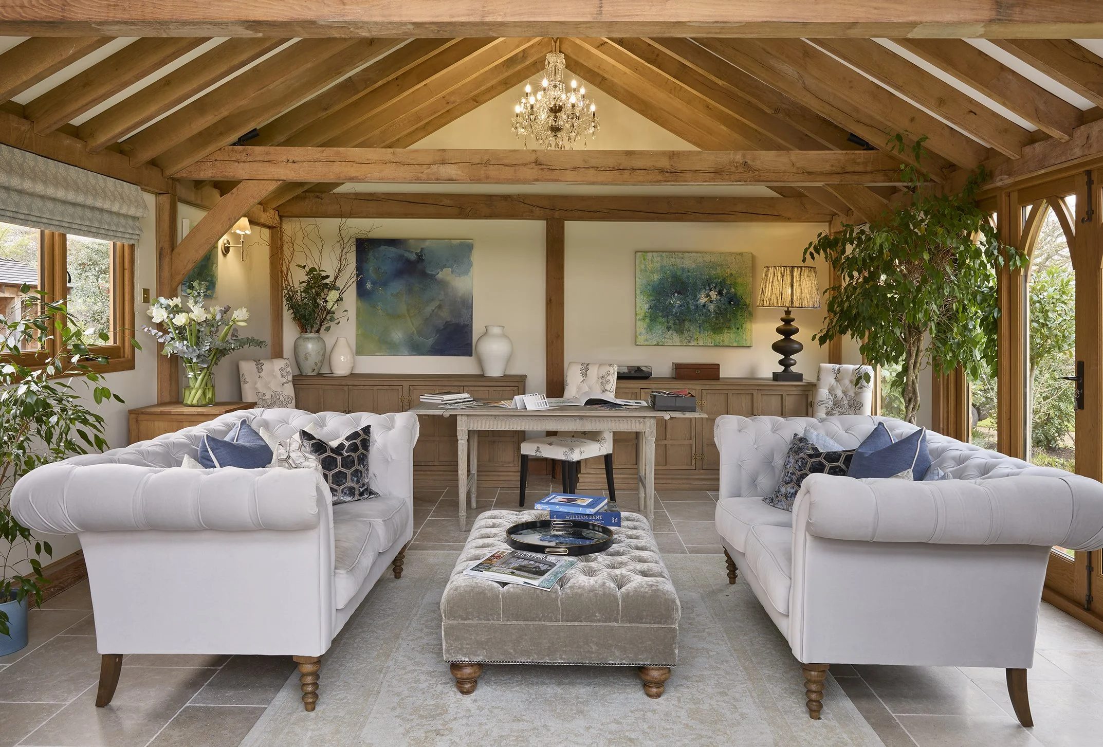 Living room with two white tufted sofas, a beige ottoman, and a wooden table, decorated with cushions and magazines, large windows, greenery, wooden beams, a chandelier, paintings, and a side table with a lamp.