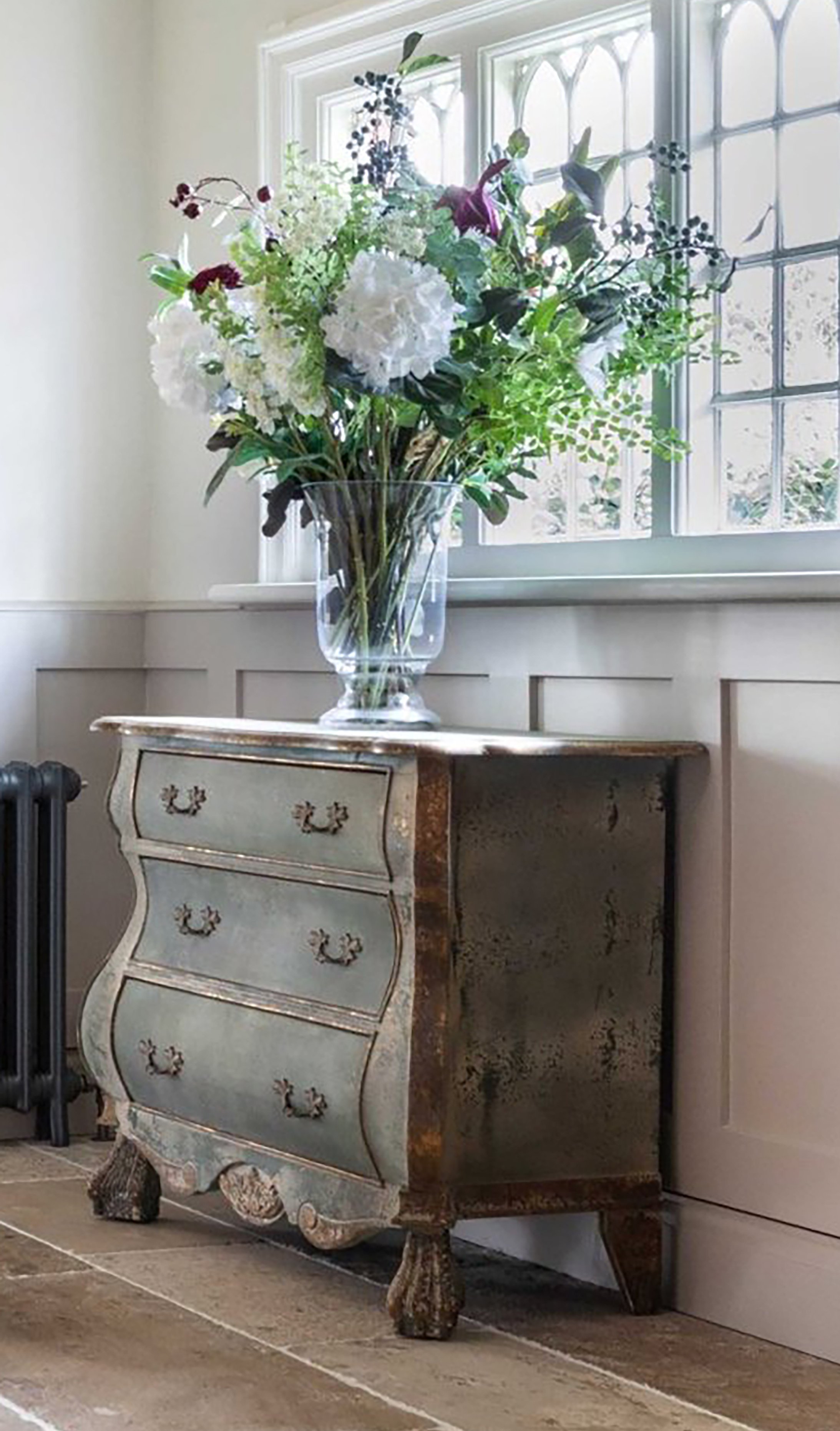 A vintage-style dresser with ornate curved lines and a distressed finish, topped with a large glass vase filled with a bouquet of assorted white, green, and deep purple flowers, set in front of a window with decorative panes.