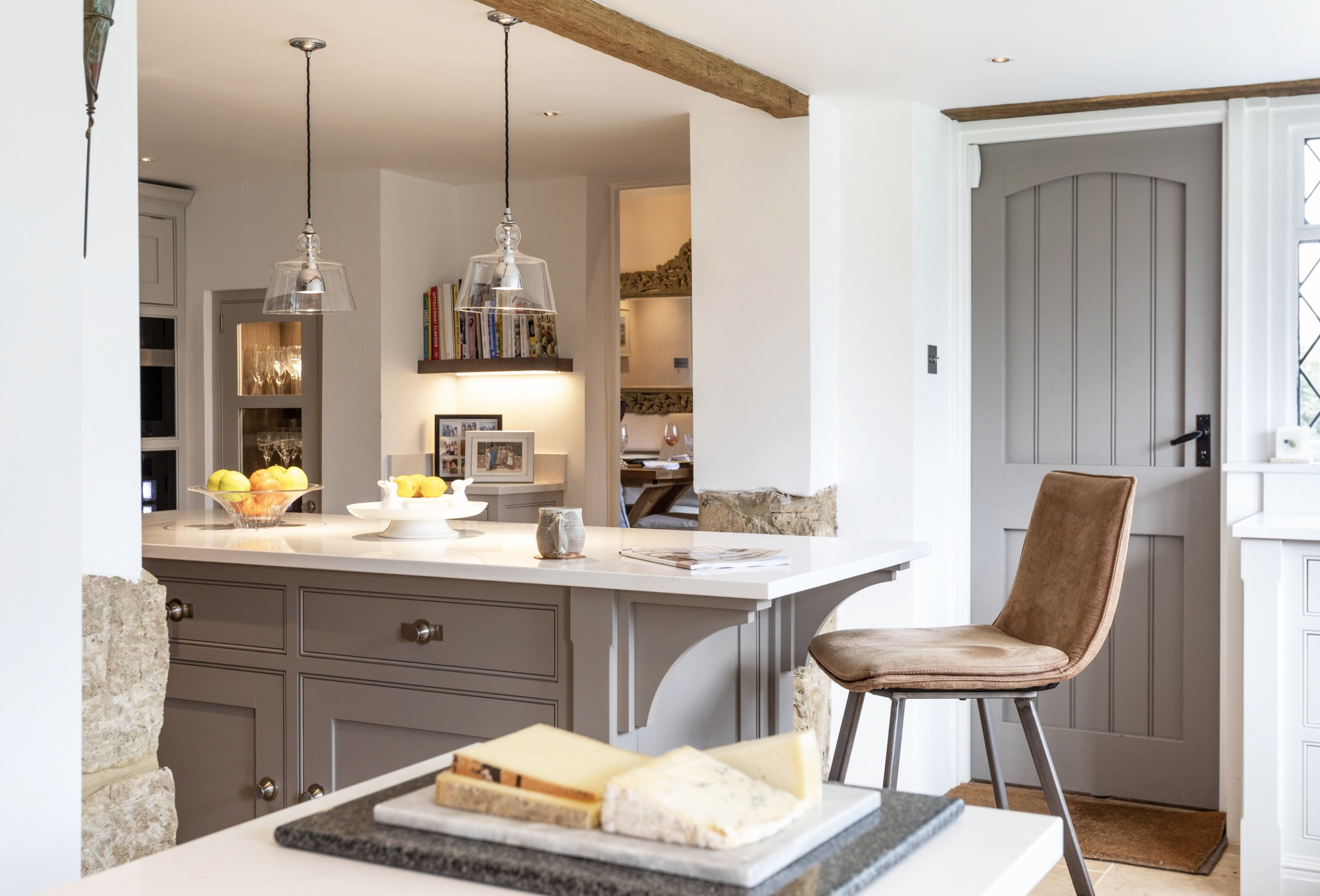 A cozy kitchen with a large white countertop, beige cabinet drawers, and a beige upholstered chair near a gray wooden door. The kitchen is decorated with fruits, photo frames, and books, with natural light coming in from the windows.