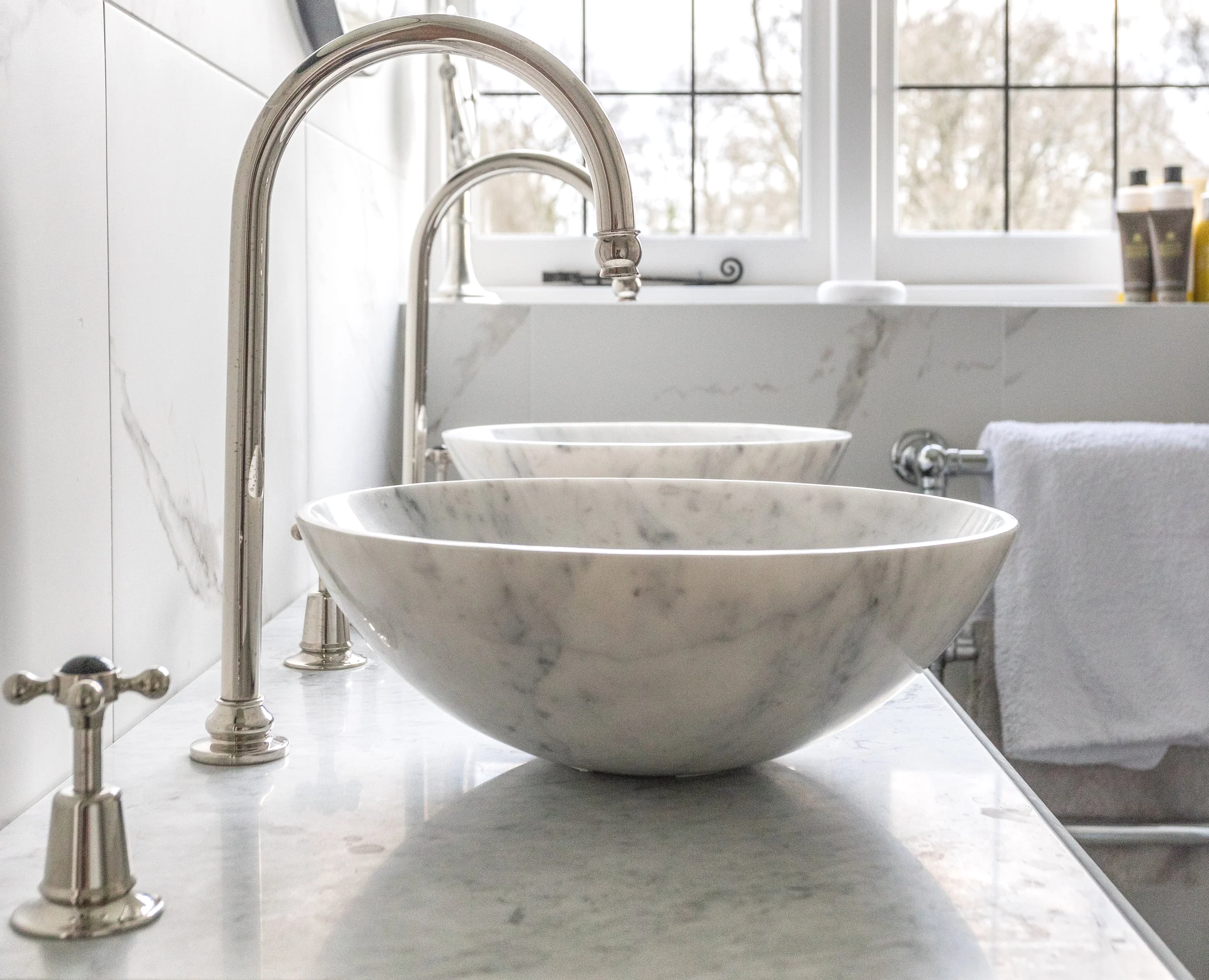 Two marble vessel sinks with chrome faucets in a bright bathroom, with a towel hanging on the side and bottles on the windowsill.