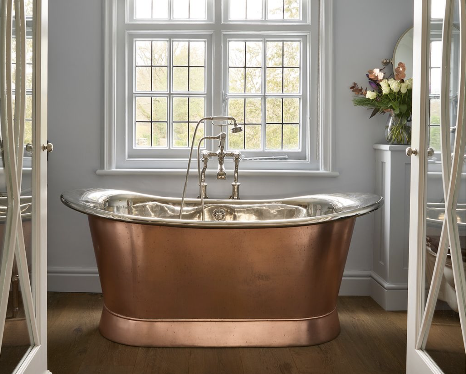 Copper bathtub in a bathroom with a large window and a bouquet of flowers in a vase.