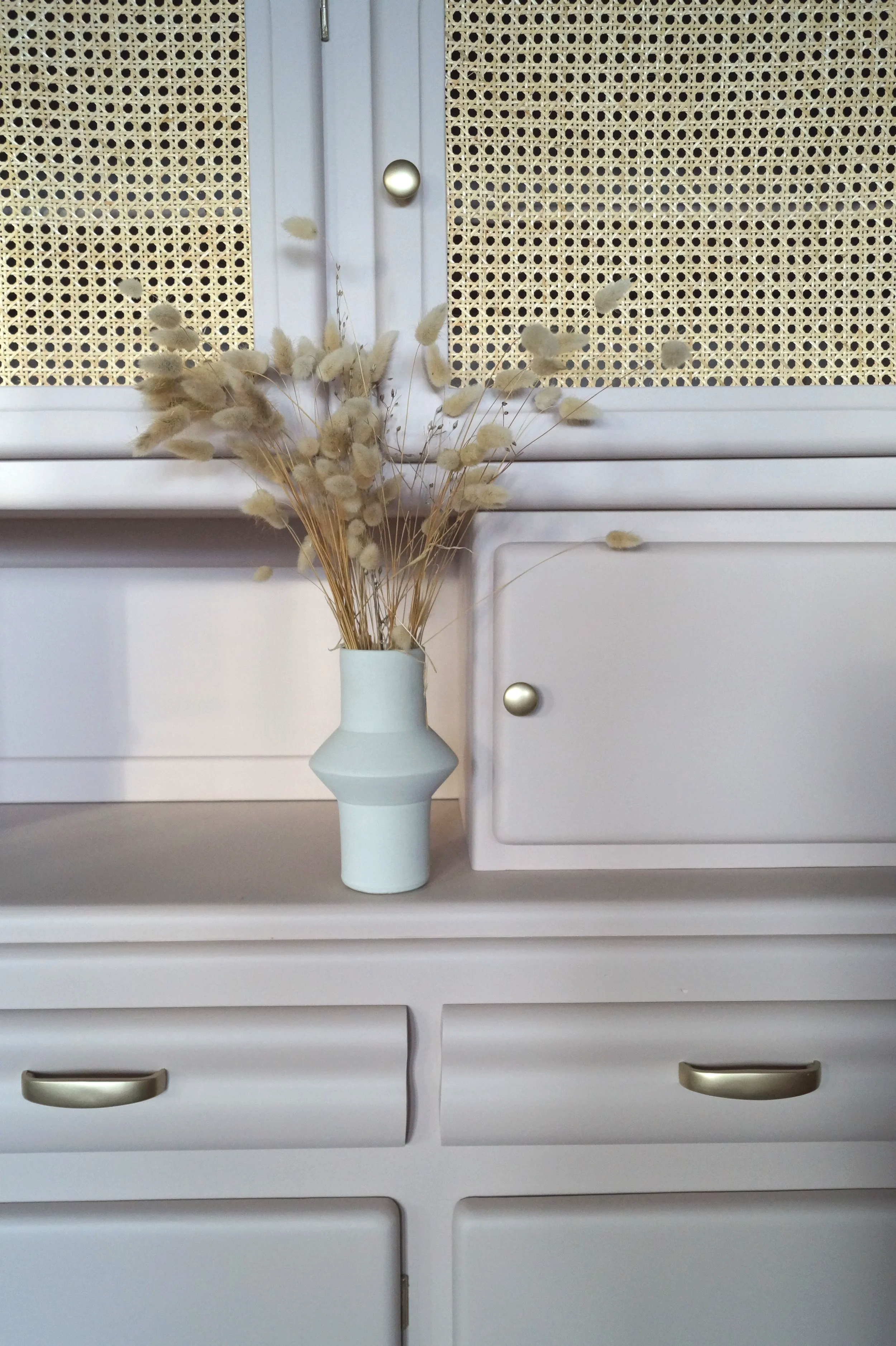 Vase blanc avec des fleurs séchées beige sur une commode blanche, en arrière-plan un meuble mado avec des portes en rotin et poignées dorées.