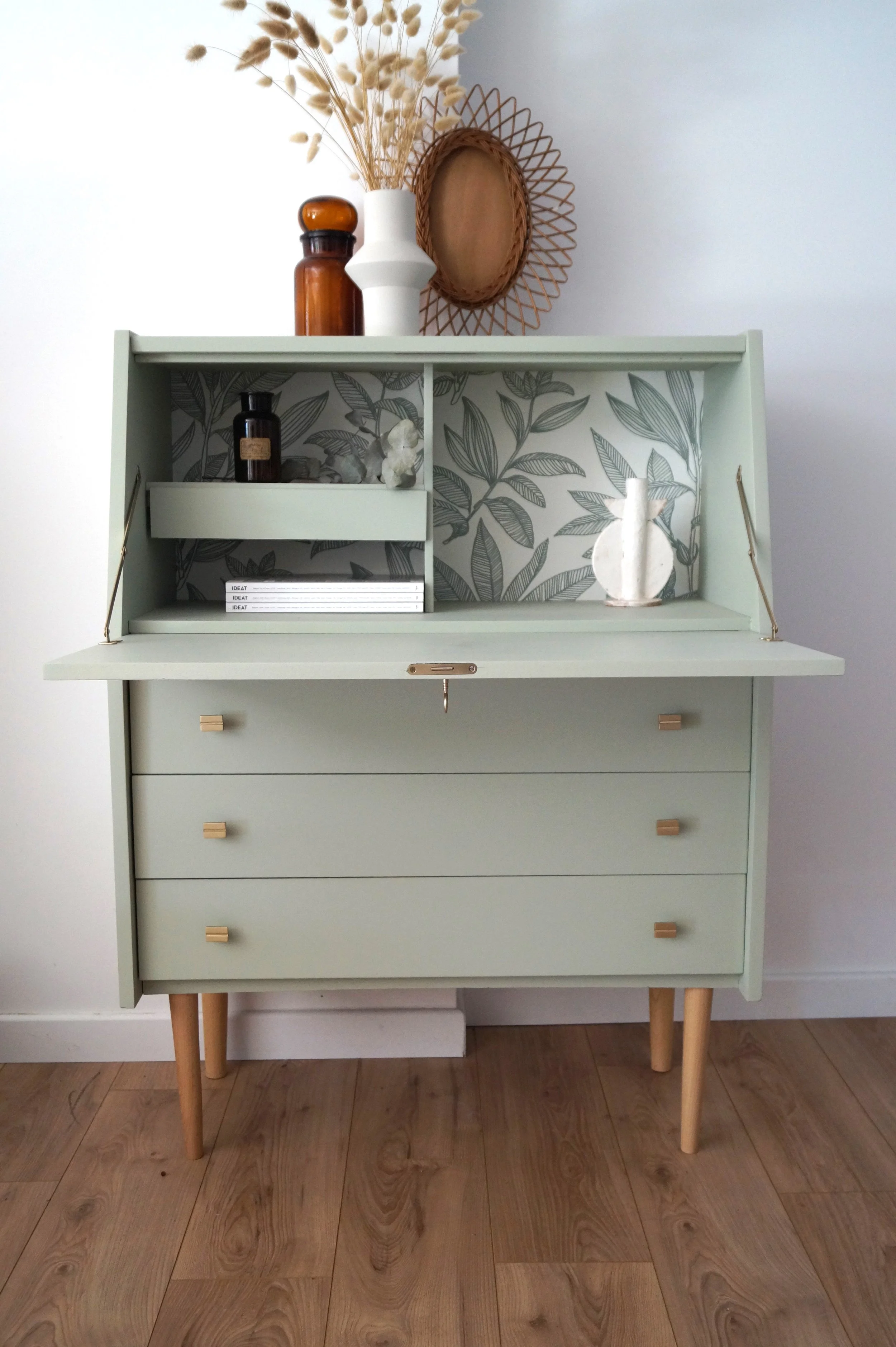 Un meuble vintage vert pastel avec trois tiroirs en bois clair, décoré avec des vases, des livres, et des objets décoratifs, avec un fond de papier peint avec motif de feuilles.