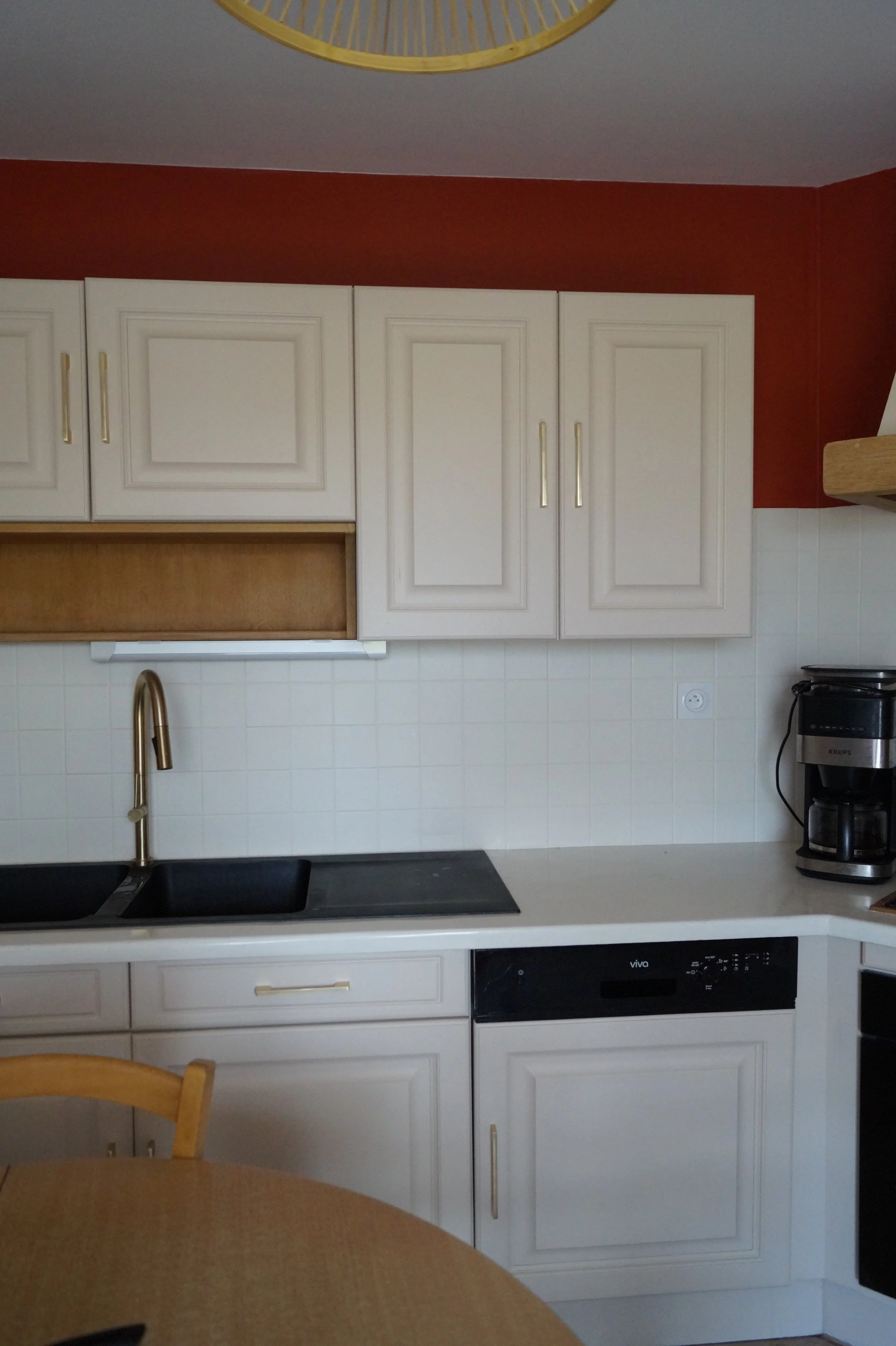 Cuisine avec un évier noir, des placards blancs avec poignées dorées, une cafetière et une table en bois.