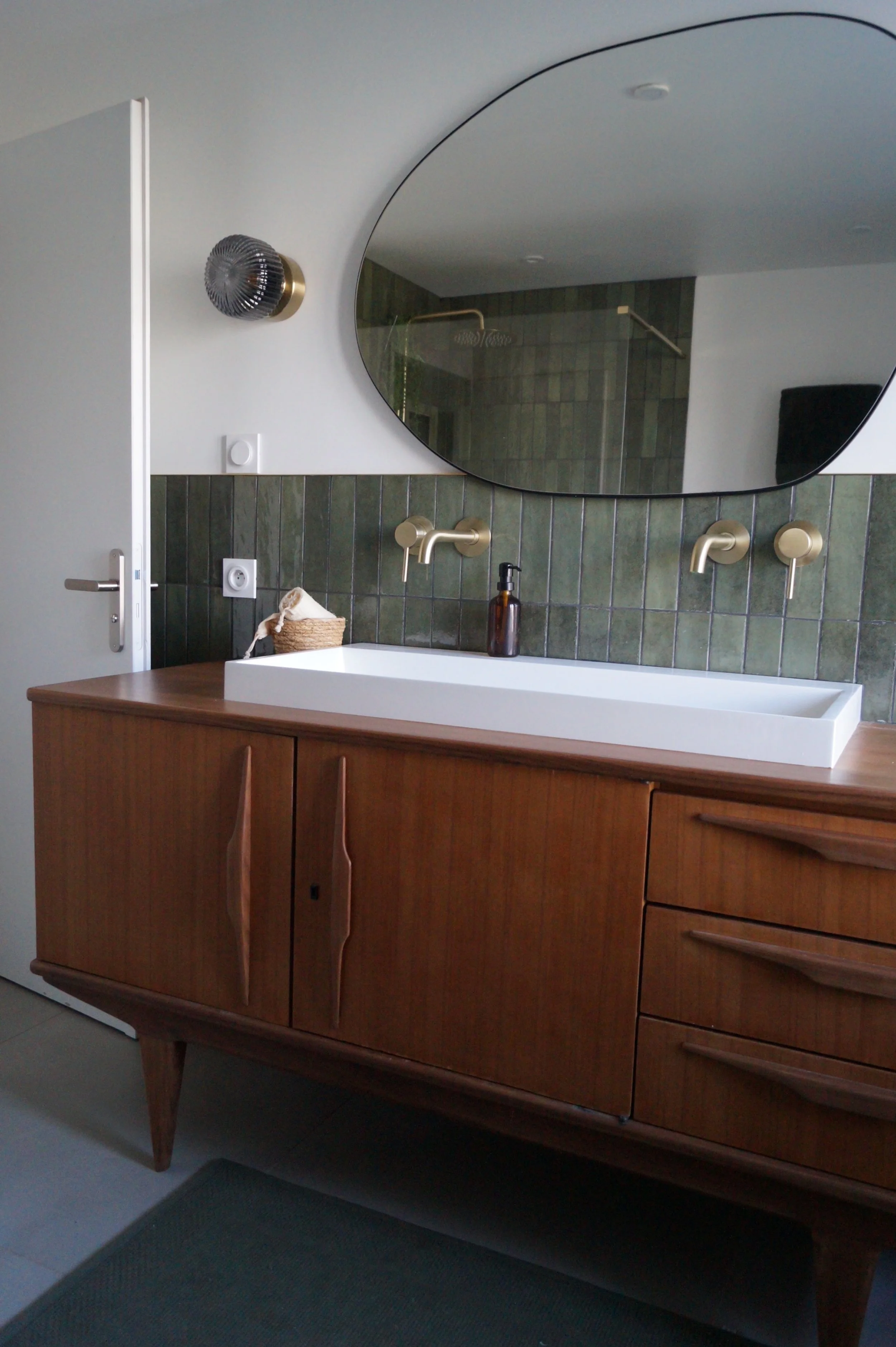 Un lavabo blanc sur une étagère en bois dans une salle de bain moderne, avec un grand miroir, un mur en carreaux verts, deux robinets dorés, une bouteille de savon et une petite panière