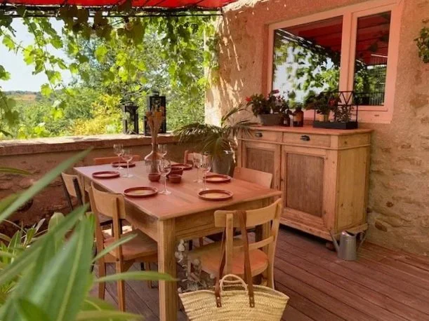 Table en bois avec six assiettes et des verres, décorée avec des plantes, à l'extérieur avec une vue sur la verdure.