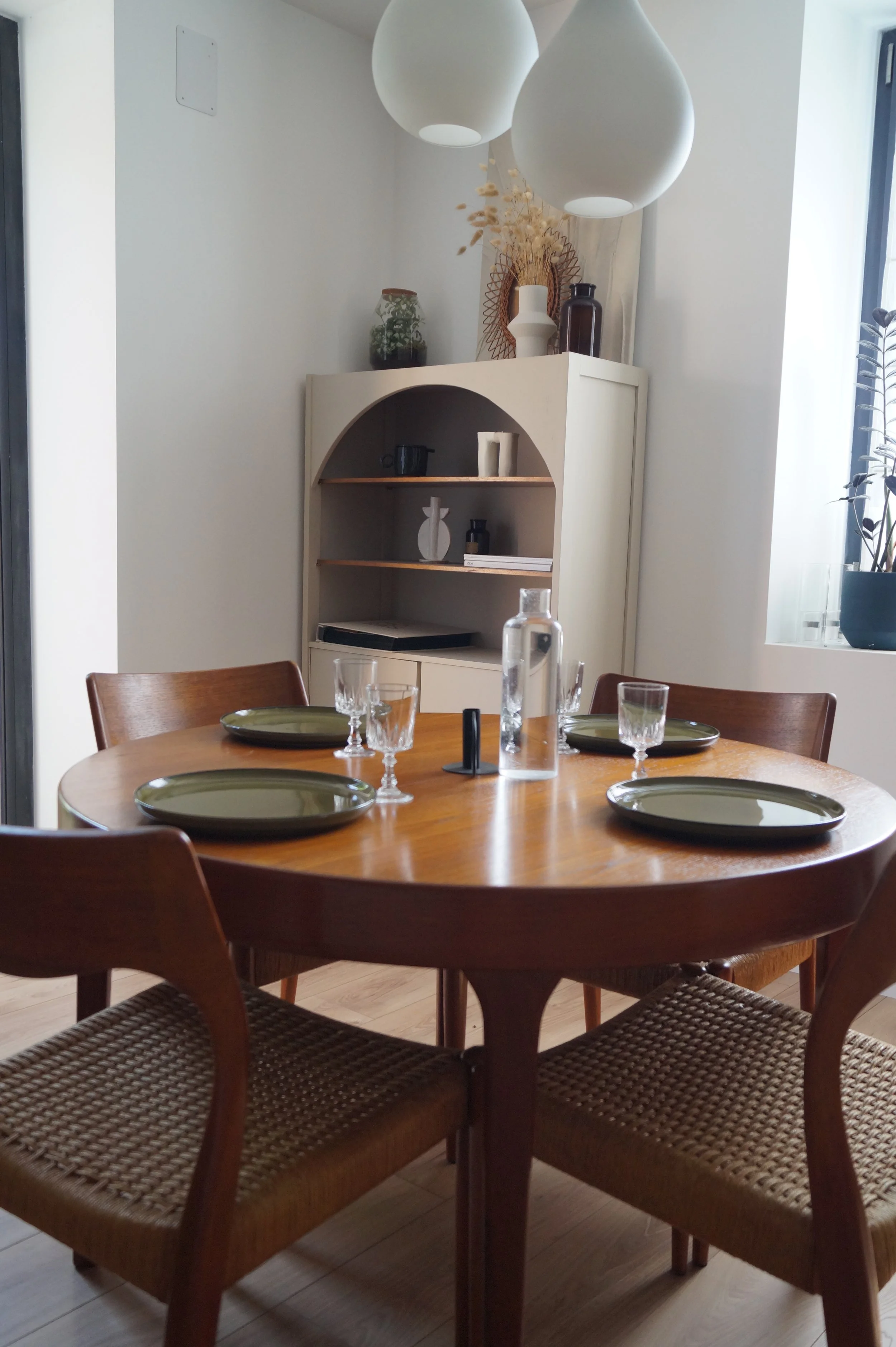 Salle à manger avec une table en bois, quatre chaises en bois avec assises en rotin, une carafe d'eau, quatre verres, quatre assiettes vertes, et une étagère blanche décorée avec des vases et des objets