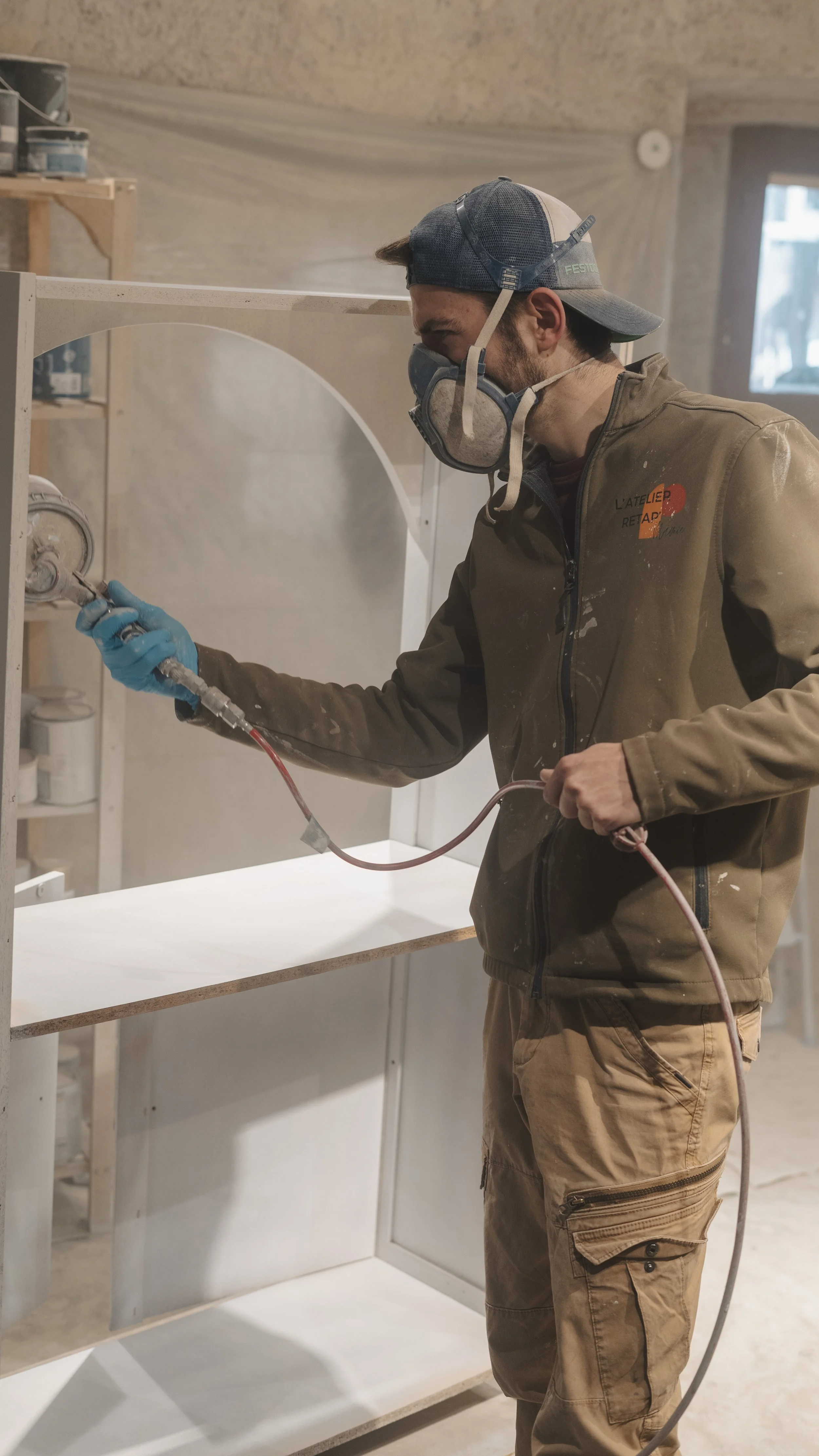 Homme portant un masque, des lunettes de protection et des gants bleus, utilisant une machine pour pulvériser de la peinture ou un autre produit sur une étagère en bois dans un atelier.