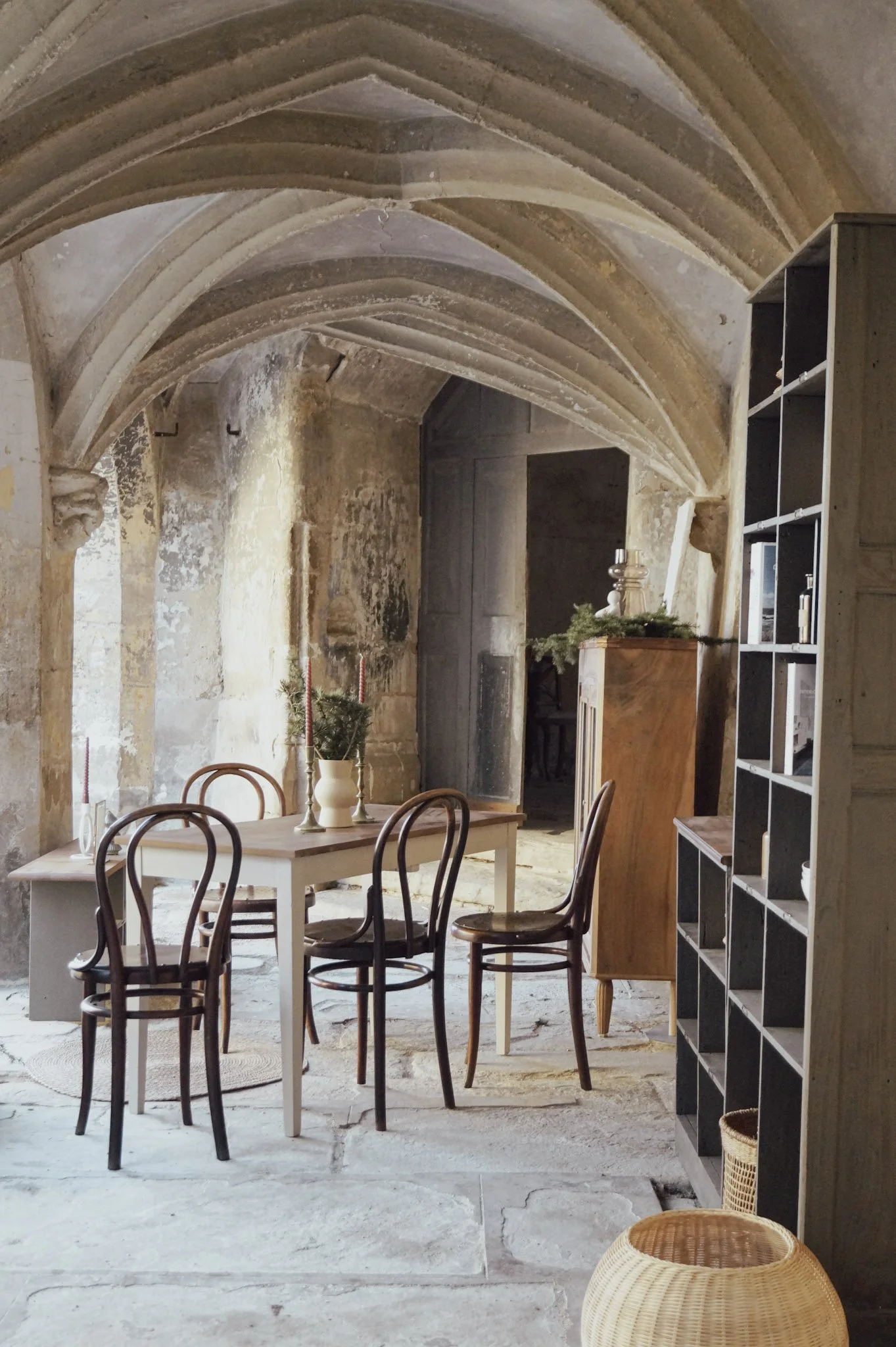 Salle à manger ancienne avec plafond en voûte en pierre, table en bois blanc, chaises en bois foncé, étagère noire avec des objets, décoration de Noël avec bougies et branches vertes.