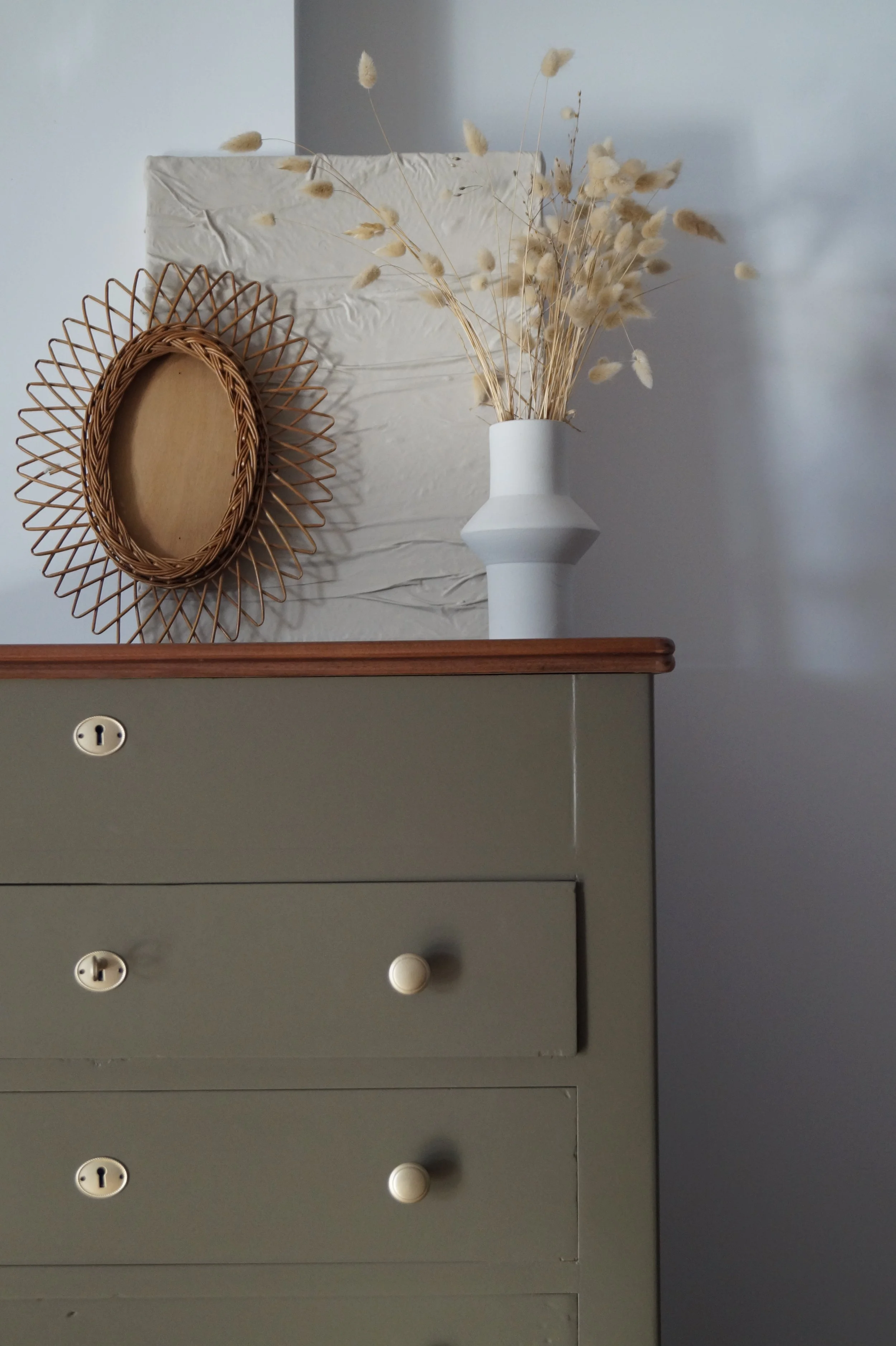 Une commode verte avec deux tiroirs, surmontée d'un vase blanc avec des fleurs sèches beiges, un miroir en rotin et un cadre en papier ondulé blanc.