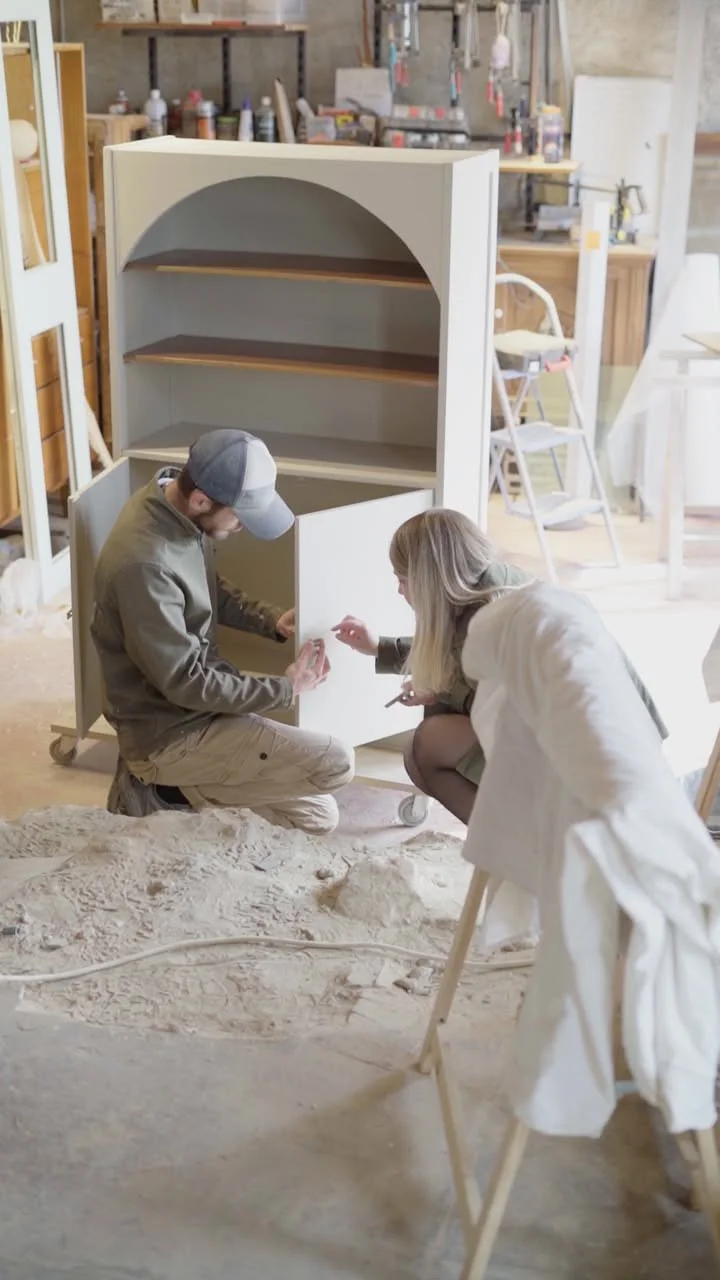 Deux personnes peignent ou assemblent un meuble en bois dans un atelier, avec des outils et matériaux en arrière-plan.