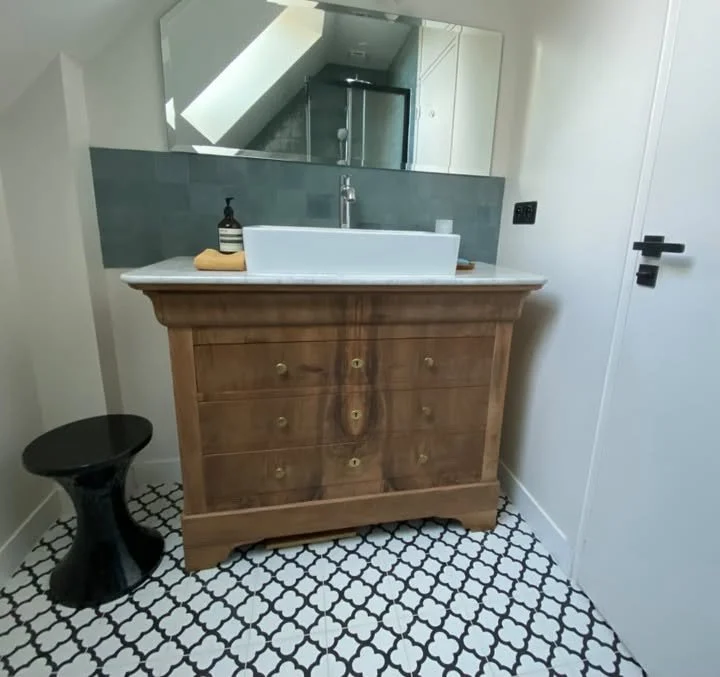 Meuble en bois avec un lavabo blanc, un miroir, un savon liquide, une serviette, un miroir au-dessus du lavabo, un petit tabouret noir, sol en carreaux noir et blanc, porte blanche à droite.