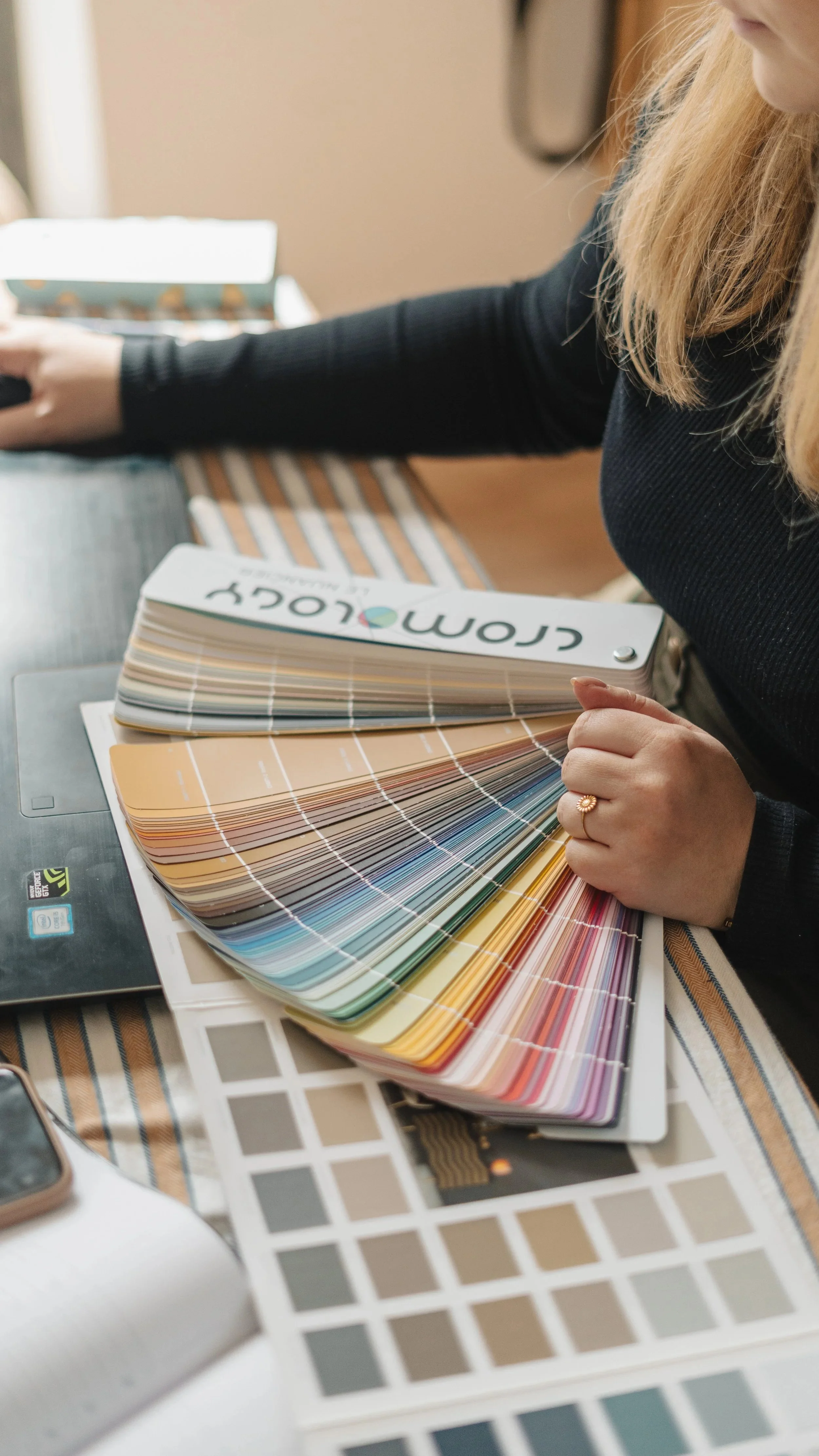 Une personne tenant une carte de couleurs dans un studio de design. Des échantillons de couleurs et des échantillons de peinture sont dispersés sur une table.