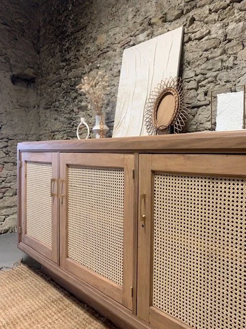 Buffet en bois avec portes en cannage, décorations sur le dessus, tableau blanc, miroir rond en lampe, deux vases, mur en pierre
