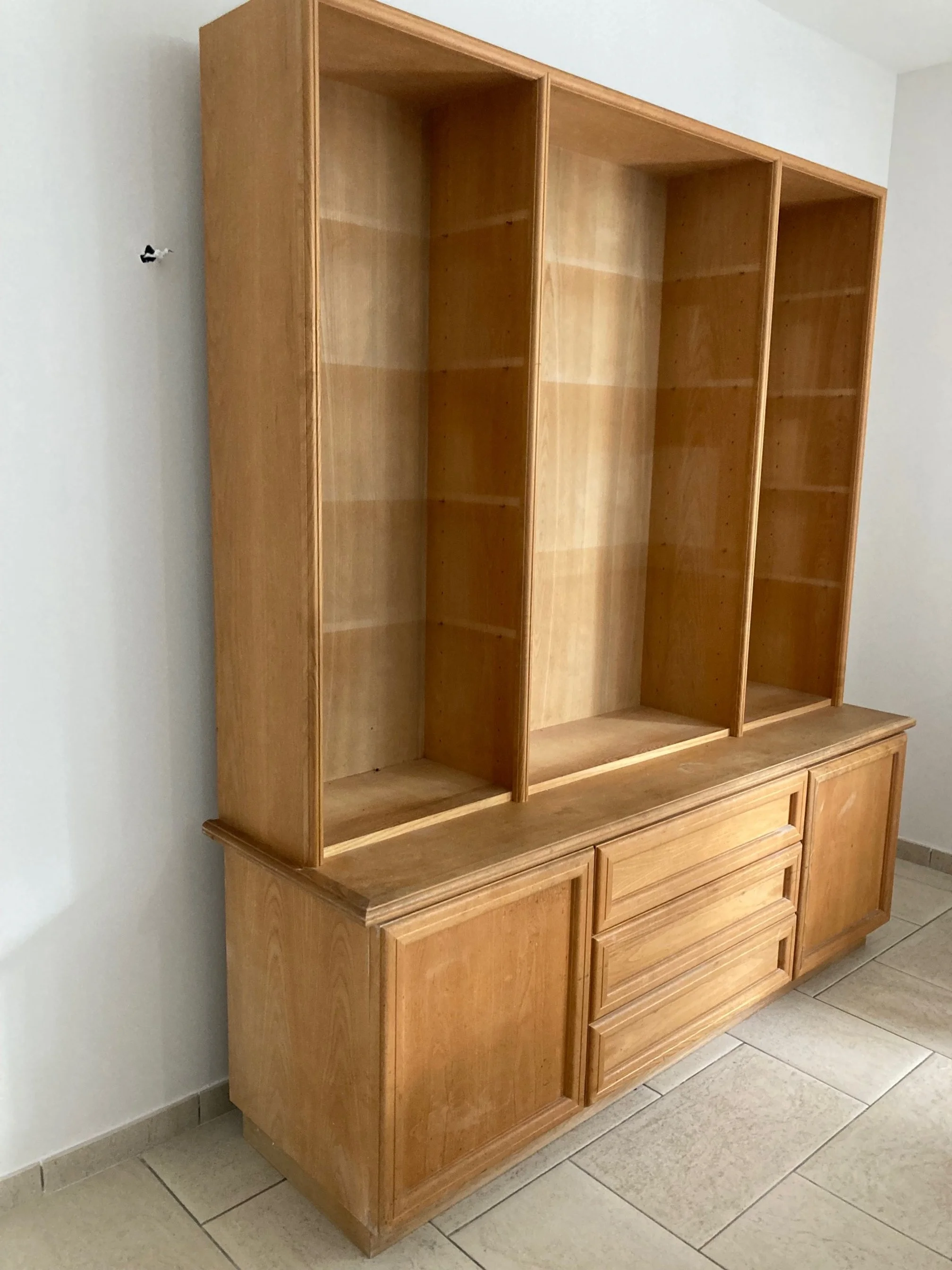 Une grande bibliothèque en bois non peinte avec des étagères vides, placée contre un mur blanc dans une pièce avec un sol en carreaux beige.