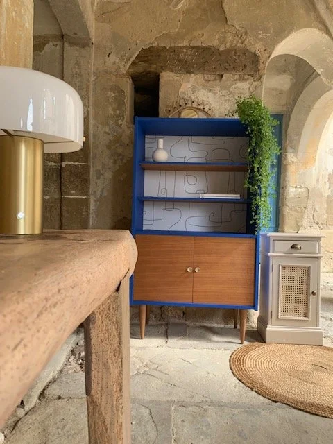 Une étagère bleue avec un décor abstrait, un vase blanc, et une plante verte en cascade dans un intérieur rustique.