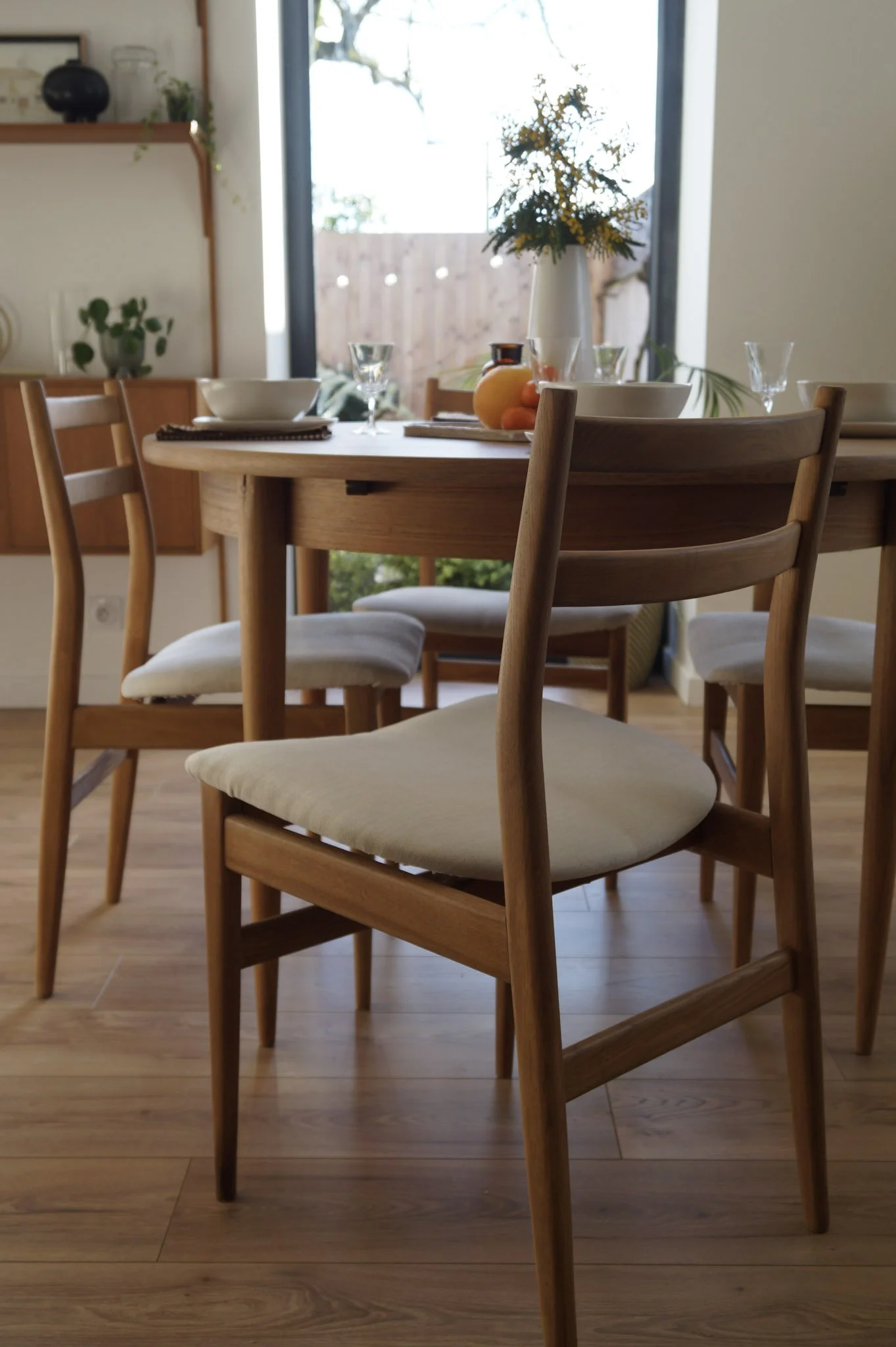 Salle à manger avec une table en bois entourée de chaises en bois avec des sièges en tissu beige, fenêtre laissant entrer la lumière naturelle, décor minimaliste avec des plantes et des objets décoratifs.