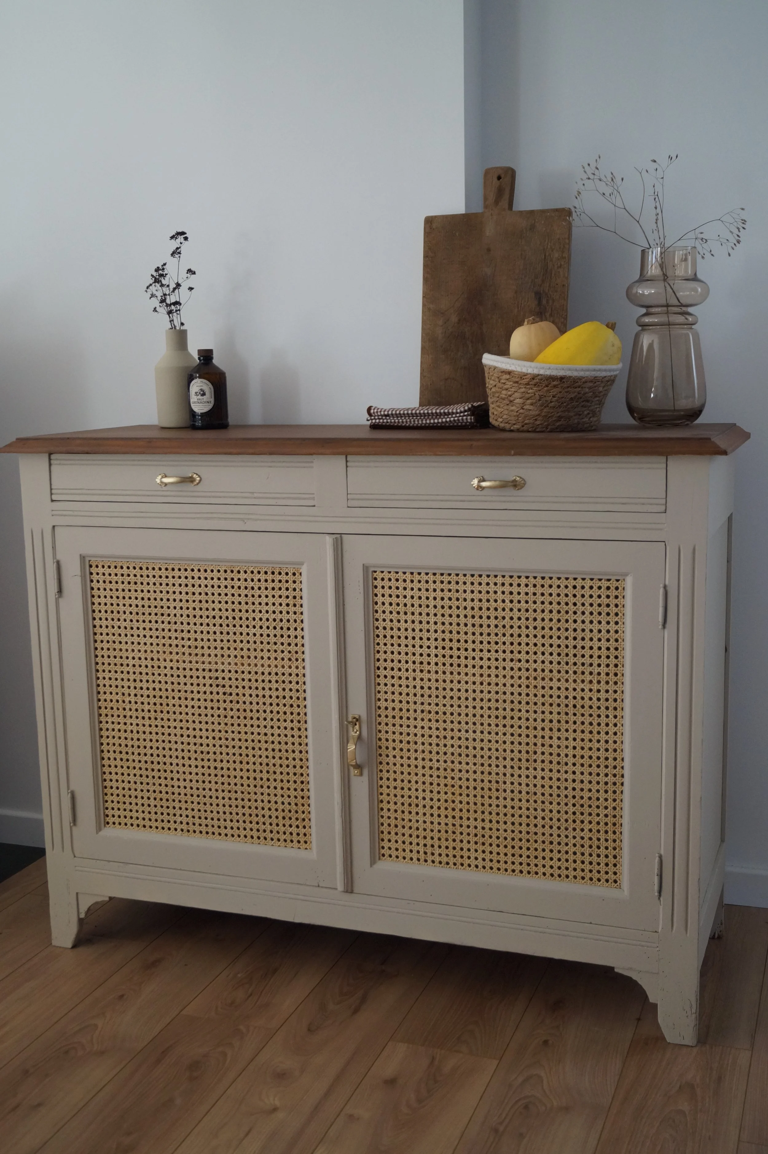 Buffet blanc avec portes en rotin, dessus en bois clair, décor avec vase de fleurs séchées, bouteille, plateau, planche à découper, paniers de fruits, vase en verre avec branches