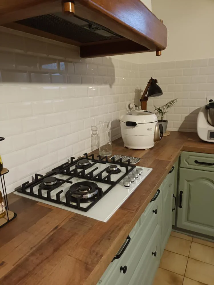 Une cuisine avec un plan de travail en bois, une gazinière à quatre feux, une lampe de bureau et un appareil de cuisson blanc. En arrière-plan, un mur avec des carreaux blancs et des éléments de cuisine.